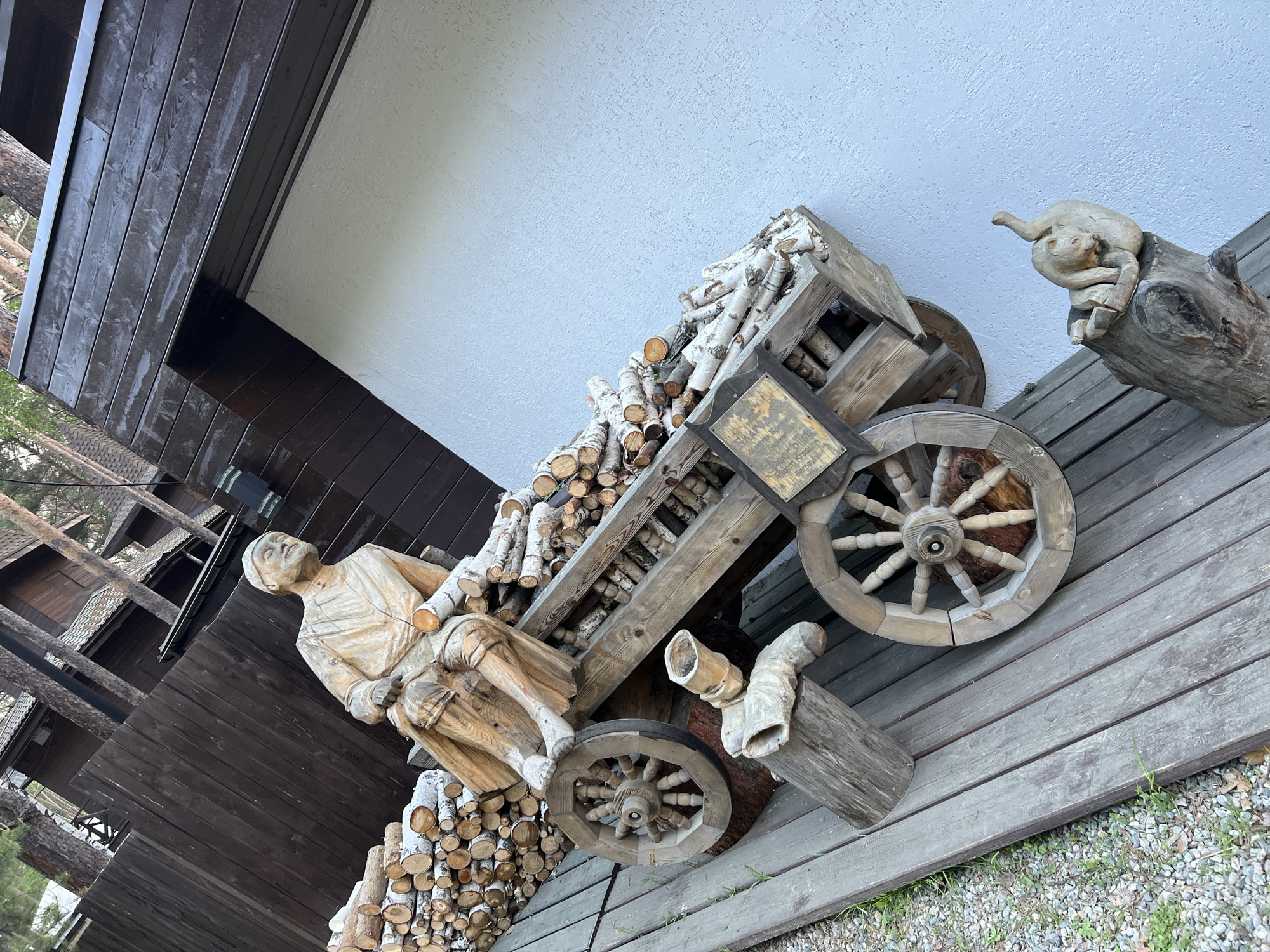 A wooden sculpture of a man sitting on a wooden cart filled with firewood is displayed outdoors. The cart has two large spoked wheels. To the left of the cart, there is a large pile of cut logs. In front of the cart, there are wooden supports for the cart's axle, and a smaller wooden sculpture of a cat resting on a log. The scene appears to be in Tal'tsy, Russia, as suggested by the architectural style of the building in the background, which features wooden siding and a dark-pitched roof. The ground is covered with a wooden walkway and gravel. The lighting indicates it is daytime.