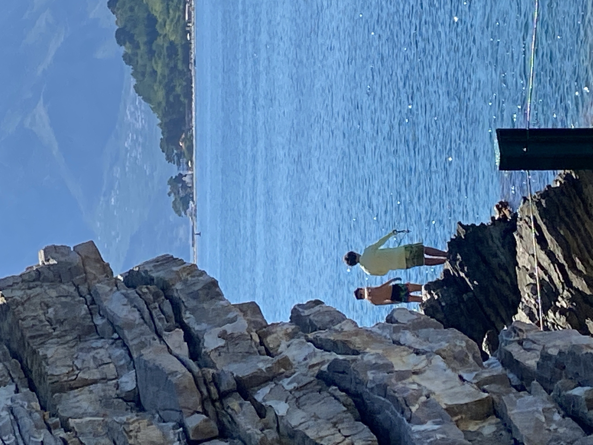 The image depicts two people standing on a rocky cliff overlooking a body of bright blue water. The person in the foreground is wearing camouflage shorts and no shirt, while the person behind them wears a light yellow shirt and camouflage shorts. They appear to be on a cliff edge, with the water below and a lush green mountainside in the distance on the left side of the frame. The sun is shining brightly, casting highlights on the water and creating shadows on the rocks. The overall scene suggests a warm, sunny day, possibly in the afternoon, given the lighting. The presence of the rocky cliff and the clear blue water are strong indicators of a coastal or Mediterranean location. Based on the provided location context of Budva, Montenegro, it's likely this scene is along the Adriatic coast, known for its dramatic cliffs and clear waters. The people appear to be engaged in an activity, possibly preparing to jump or enjoying the view from their perch. There is no visible text in the image.