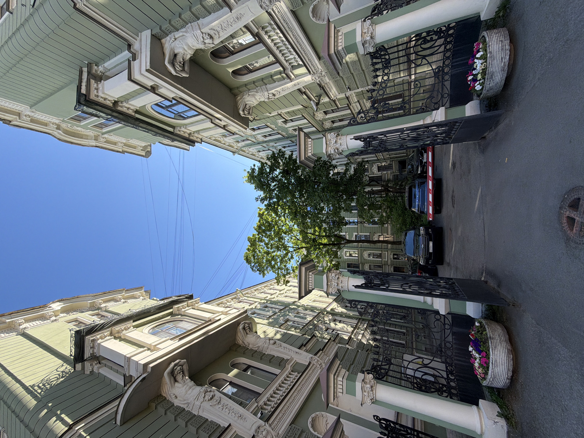 This image depicts a low-angle view looking into a courtyard entrance framed by two ornate buildings in Saint Petersburg, Russia. The multi-story buildings, painted a light sage green with white architectural trim, feature detailed facades including pilasters, cornices, balustrades, and sculpted male figures (atlantes) supporting balconies on the upper levels. A pair of large, open black wrought-iron gates, supported by light green and white fluted columns with ornate capitals, defines the entrance. Immediately inside the gates, two oval concrete planters contain colorful mixed flowers, predominantly pink, white, purple, and yellow.
The paved courtyard beyond the gates contains two dark-colored vehicles: a Toyota Land Cruiser with Russian license plate X206XT 178, and a Volvo with Russian license plate K501AK 178 parked behind a partially lowered red and white striped boom gate. Green deciduous trees with full foliage are visible deeper within the courtyard. Overhead, several thin wires traverse a clear, bright blue sky. Strong sunlight casts distinct shadows on the building facades. A circular metal manhole cover is visible on the asphalt in the foreground. No individuals are present in the scene.