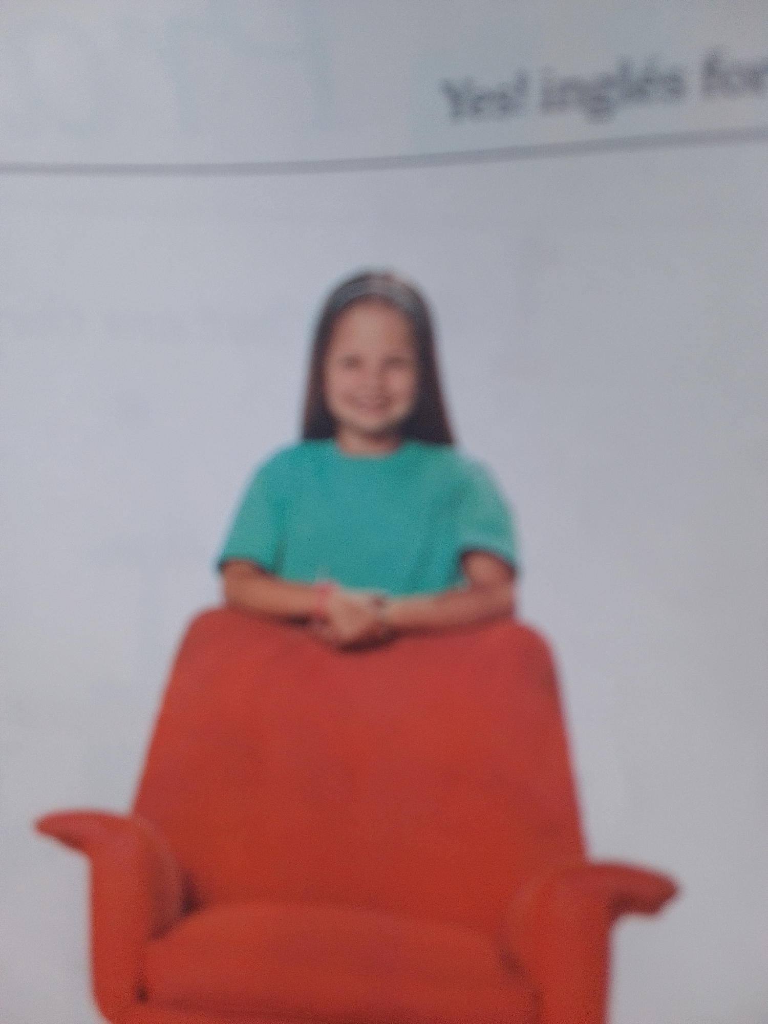 A young girl with brown hair, wearing a teal shirt and a pink bracelet, sits in a bright orange armchair. Her hands are clasped in her lap, and she has a slight smile on her face. The background is a plain, light gray, suggesting an indoor setting. There is text visible in the upper right corner of the image, reading "Yes! inglés for". The image is slightly out of focus, giving it a soft, almost dreamlike quality. The overall impression is one of a candid moment captured, perhaps during a lesson or a photo shoot. The setting of La Tagua, Venezuela, is not visually evident in the image itself, but it provides a geographical context for the photograph.