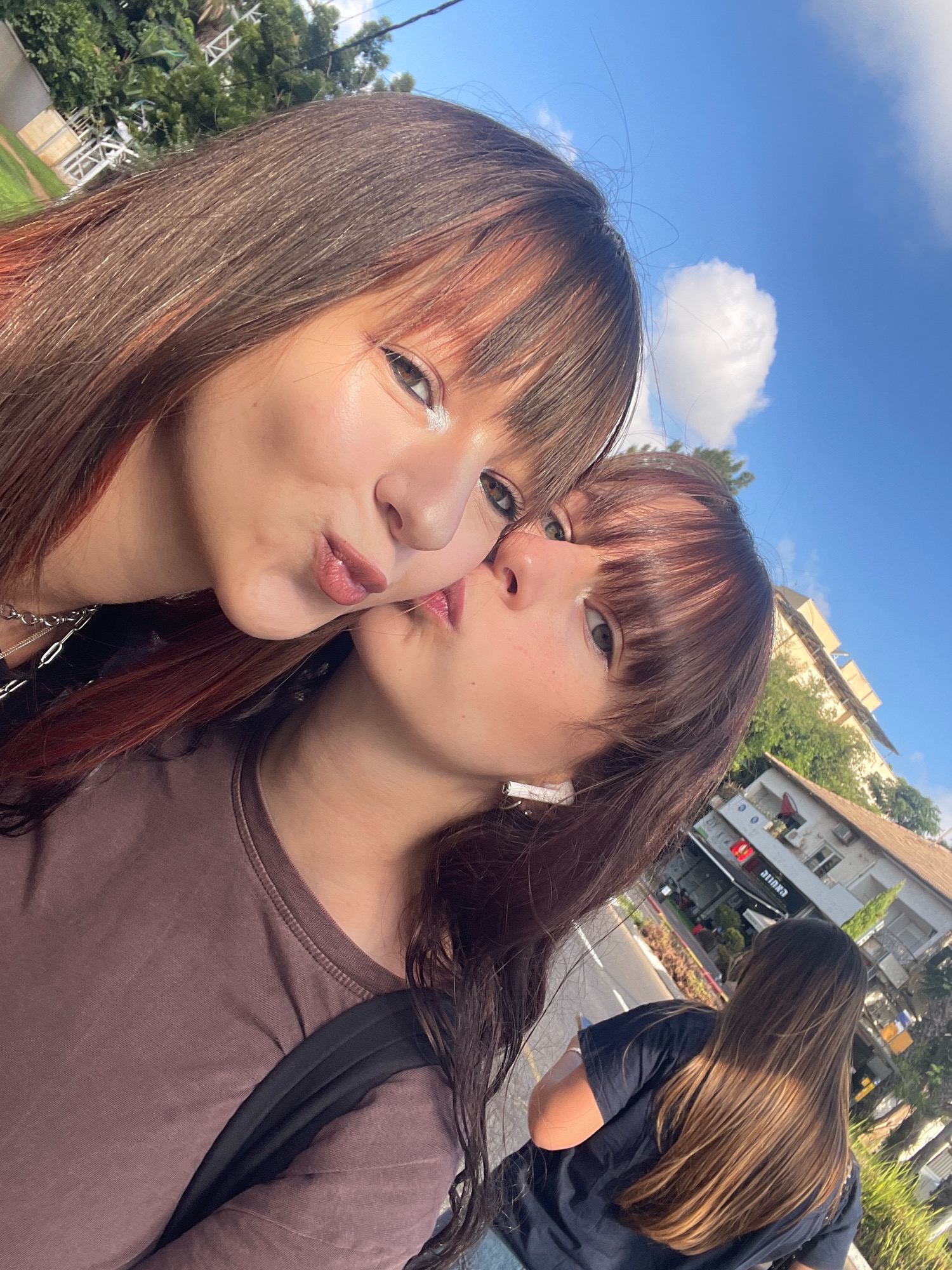 In Raanana, Israel, two young women are taking a selfie outdoors. The woman in the foreground on the left is looking at the camera with her lips pursed, as if leaning in for a kiss. Her hair is long and brown with reddish highlights. She is wearing a dark shirt and a silver chain necklace. The woman on the right is leaning her head towards the woman on the left, with her lips touching the other woman's cheek. She also has long, brown hair and is wearing a brown t-shirt and white earbuds. In the background, a third person with long brown hair is walking away from the camera. They are wearing a dark t-shirt. The scene appears to be a street with buildings and trees, under a bright blue sky with some clouds. The architecture visible suggests a Mediterranean setting.