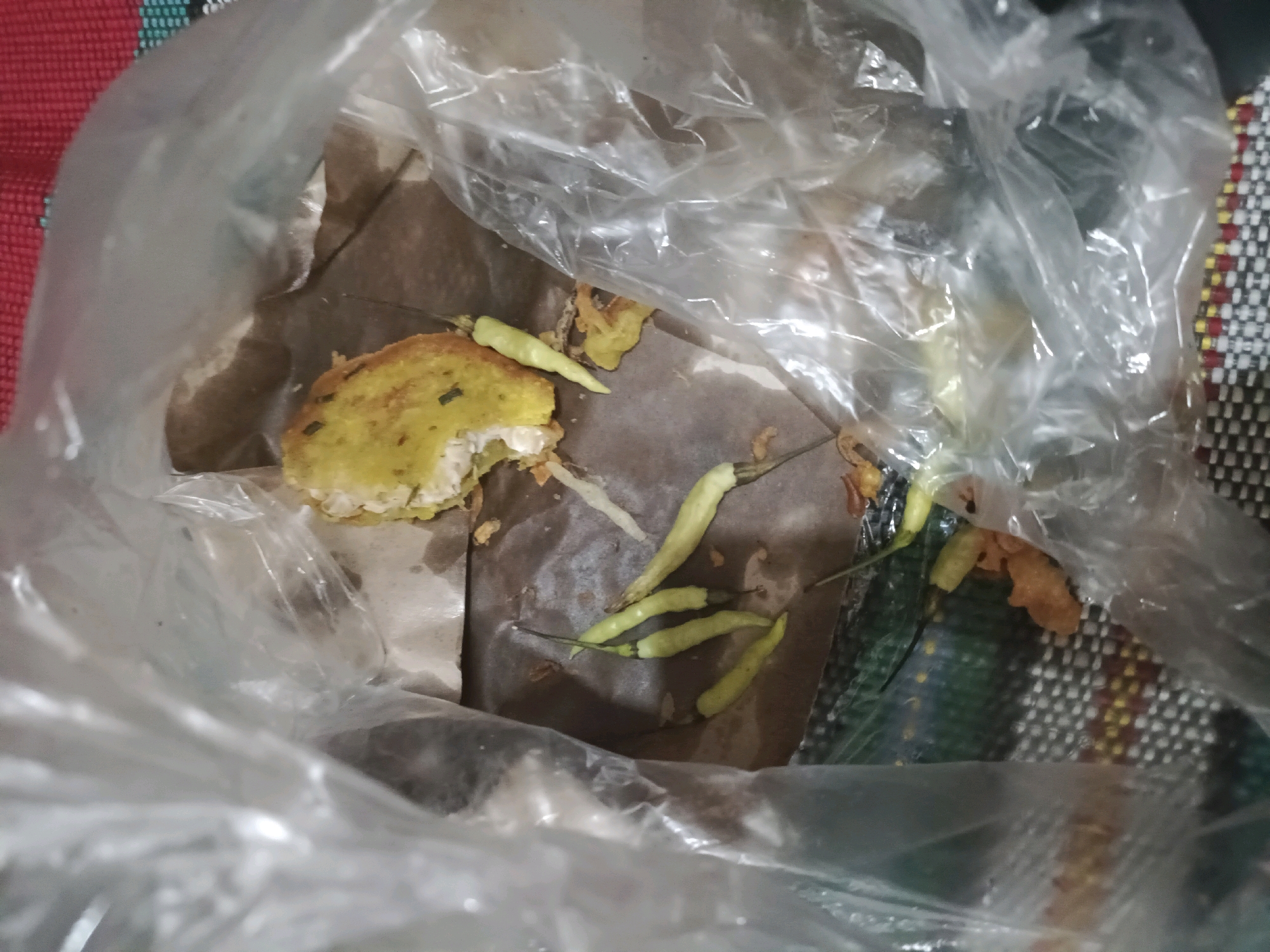 The image shows a close-up view of food scraps inside a clear plastic bag. The food appears to be pieces of a fried fritter and several small green chilies. The fritter is golden brown with some green flecks, and a bite has been taken out of it. The chilies are whole and still attached to their stems. The food is placed on a piece of brown paper. The plastic bag is partially open, and the background is a red and white patterned fabric, possibly a placemat or tablecloth. The overall scene suggests discarded food. The image does not contain any information to determine its location in the city of Rancang Kencono, Indonesia.