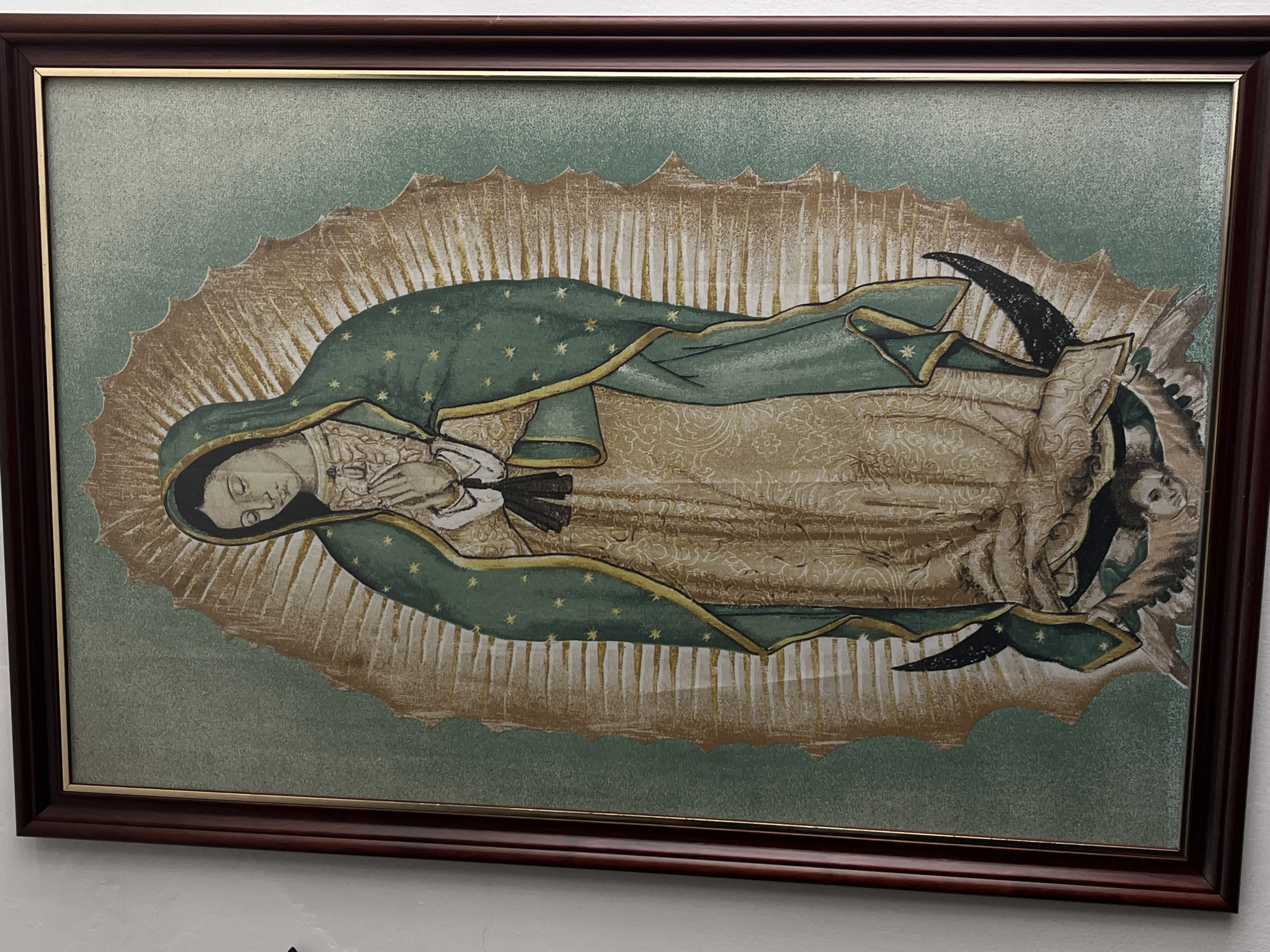 A framed image depicting the Virgin of Guadalupe hangs on a white wall. The image features a depiction of the Virgin Mary in a green cloak adorned with stars. The background of the image is a light green with gold rays emanating from the central figure. The frame is dark brown with a thin gold inner border. The location is Unknown, Unknown.