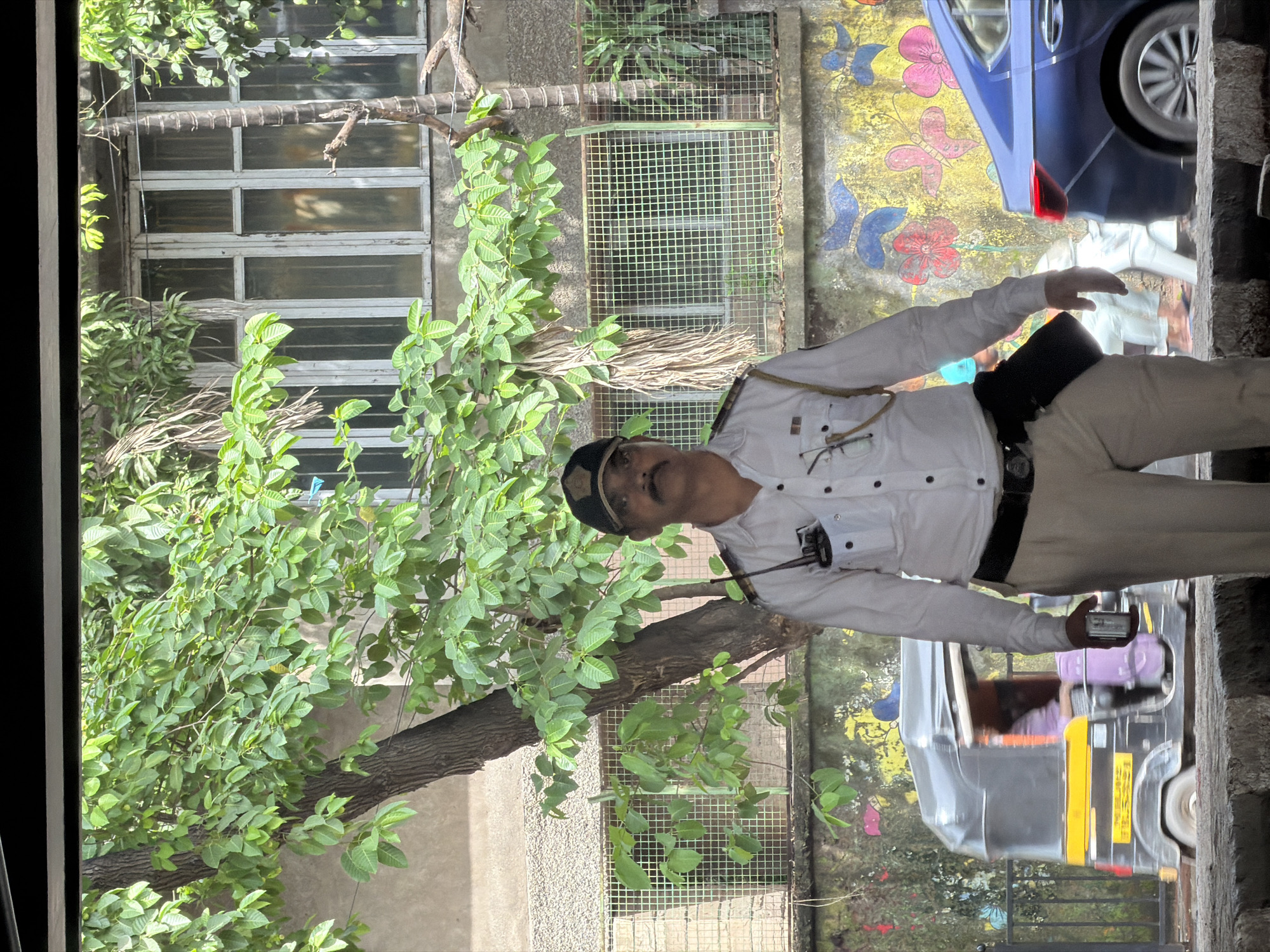 A traffic police officer stands in the foreground, facing forward, in a well-lit outdoor urban environment. The officer is wearing a white long-sleeved shirt with epaulets, a dark cap, tan trousers, and a black belt with a black holster attached. They are holding a small silver device in their right hand. Behind the officer, a tree with green leaves occupies a significant portion of the frame. Further in the background, a building with multi-paned windows is visible. The wall behind the officer is adorned with colorful graffiti depicting butterflies and flowers. To the left, a yellow and black auto-rickshaw is partially visible, and to the right, the rear of a blue car is present. The overall scene suggests a typical street in Mumbai, India, likely a busy intersection or thoroughfare.