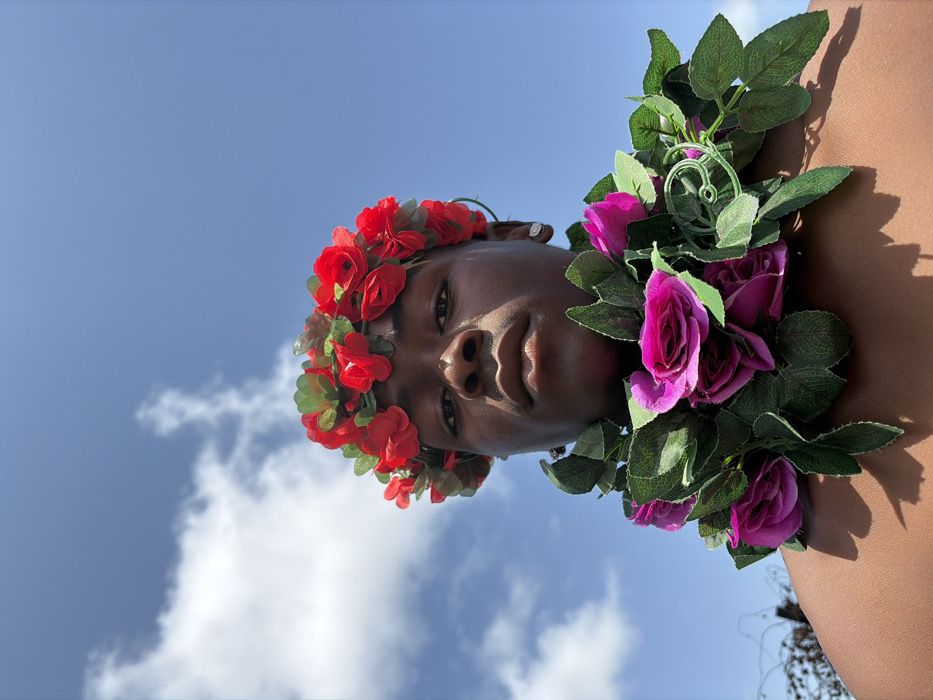 The image captures a dark-skinned individual, appearing male, positioned outdoors against a clear blue sky dotted with white clouds in Igboefon, Nigeria. The individual is framed from the upper chest, looking directly into the camera with a neutral expression. A circlet of red flowers and green leaves adorns the head, and a garland of purple flowers and green leaves is draped around the neck and shoulders. A single, small, reflective stud earring is visible in the subject's right ear. Direct sunlight illuminates the scene, casting shadows on the right side of the face and the left shoulder. A blurred, dark, indeterminate object is partially visible on the far left background.