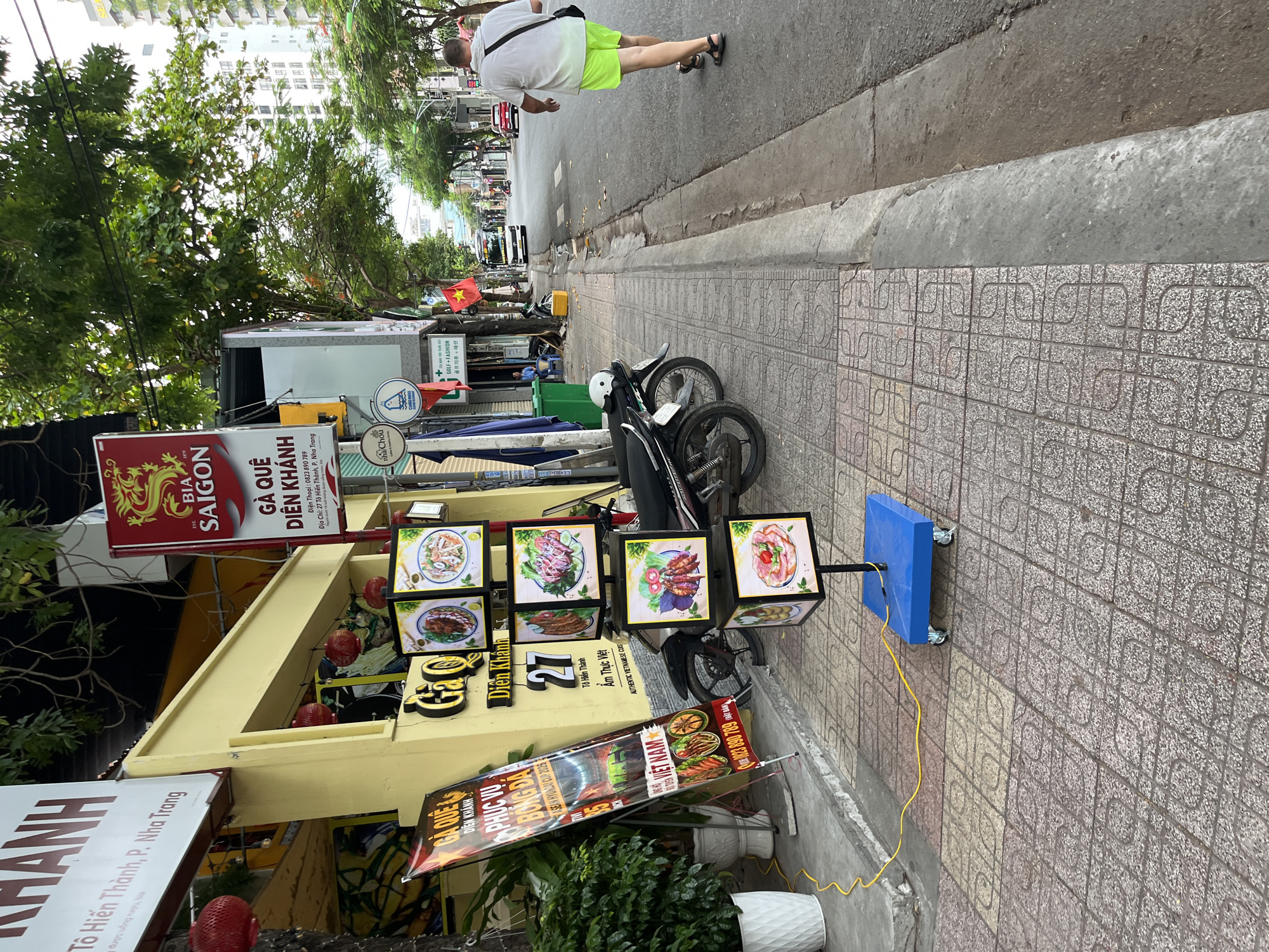 A street-level, eye-level, medium shot captures a scene on a sidewalk in Nha Trang, Vietnam. The pavement is tiled with a mosaic of light brown and grey square tiles. A person walks away from the camera on the right side of the frame, wearing a white t-shirt and bright green shorts. They are carrying a dark bag over their left shoulder and wearing dark sandals. In the background, a street stretches into the distance with traffic, including a white bus and a red car.
On the left side of the image, a yellow building forms the backdrop. Several red paper lanterns hang from the eaves of the building. A tall, narrow banner with Vietnamese text and images of food is propped up on the sidewalk. Next to it, a display unit with four square frames, each showcasing an image of food, is mounted on a blue wheeled base. The display unit is powered by a yellow electrical cord. Two motorcycles are parked on the sidewalk.
The signage on the yellow building includes the words "BIA SAIGON" and "GA QUE DIEN KHANH." Another sign indicates "27 TO Hien Thanh, P. Nha Trang". Further down the sidewalk, more signage can be seen, including flags and what appear to be shop entrances. Green trees with lush foliage overhang the scene. The lighting suggests it is daytime, with overcast or diffused sunlight.