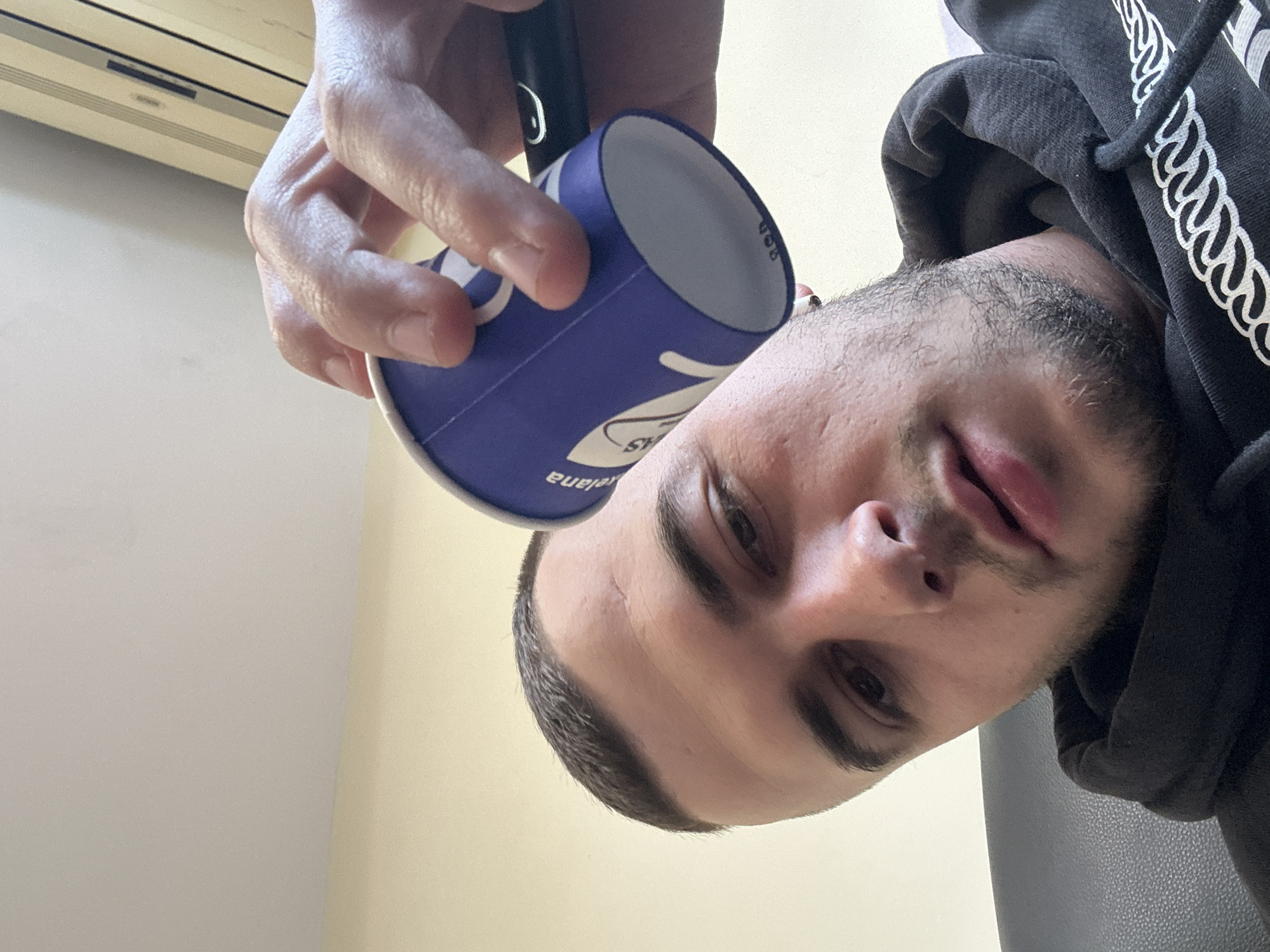 A man is holding a blue and white paper cup near his face. He is wearing a black hoodie. The location appears to be indoors, based on the wall and air conditioning unit in the background. The location in the city of Plovdiv, Bulgaria cannot be determined.