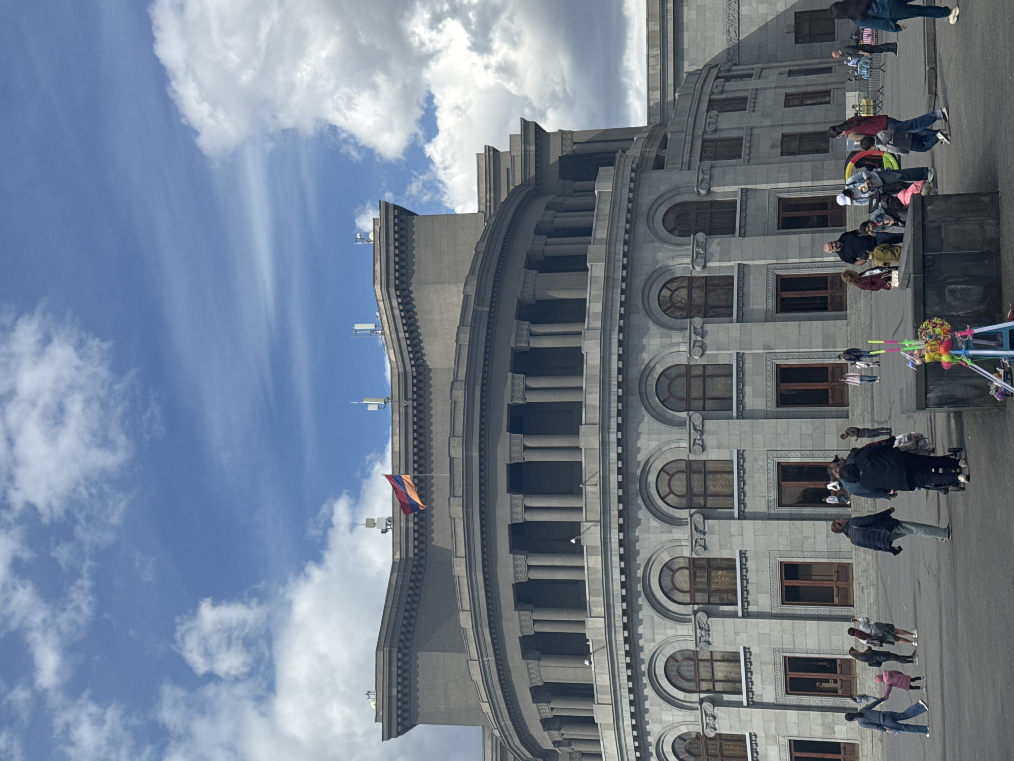 The image captures the Government House, located in Republic Square in Yerevan, Armenia. The building is a large, light-colored stone structure with multiple stories, characterized by arched windows and columns. A flag of Armenia is visible atop the building. A crowd of people are standing and walking outside the building. The sky is blue with scattered clouds.