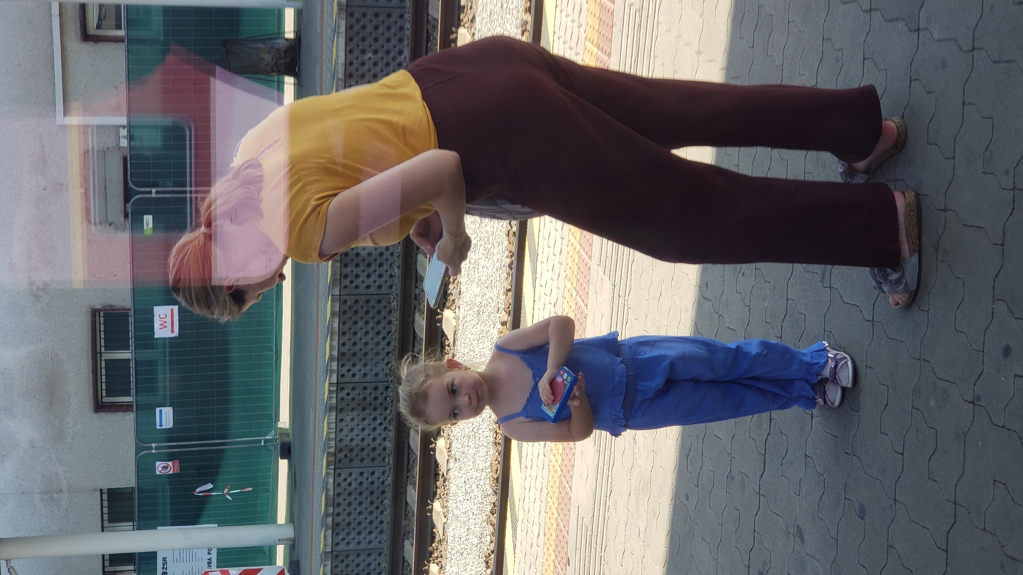 A woman in a yellow shirt and maroon pants stands on a train station platform, holding a smartphone. Beside her, a young girl in a blue jumpsuit holds a toy. The platform is paved with interlocking concrete blocks. In the background, train tracks are visible, as well as a fence and a building. The image appears to have been taken at the train station in Trenčín, Slovakia.