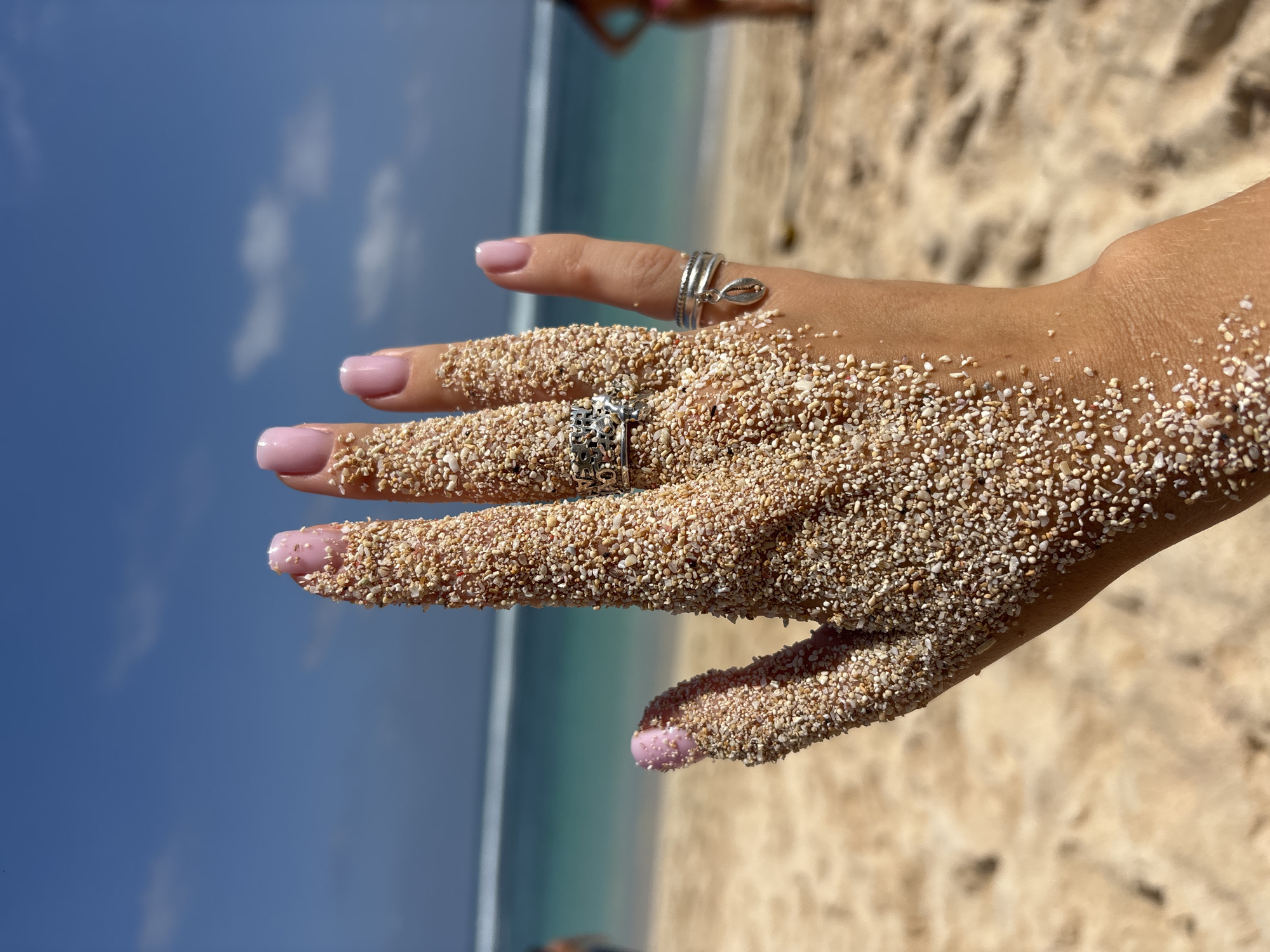 In an outdoor, natural environment, a close-up shot focuses on a left hand covered in light brown sand. The hand is held up, palm facing away from the camera, with fingers extended. The sand adheres to the skin and fingernails, which are painted a light pink color. The hand is adorned with three silver rings. One ring on the middle finger is intricate and appears to have small stones. Another ring on the ring finger is a band with a small seashell charm. A third ring is on the index finger.
The background of the image depicts a beach scene under a bright blue sky with scattered white clouds. The ocean, with turquoise water and a white foamy wave line, is visible behind the hand. The sand of the beach stretches out in front and to the sides of the hand, showing subtle footprints. Two individuals are partially visible and out of focus in the distance, on the beach near the water's edge. The overall lighting suggests a sunny day. The location is described as Unknown, Unknown.