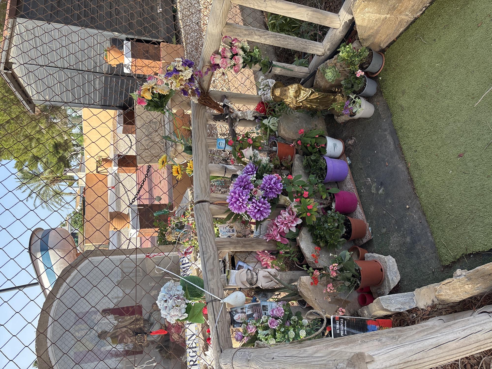 The image depicts an outdoor shrine, possibly a roadside or memorial, adorned with numerous artificial and natural flowers, potted plants, and religious figurines. A golden statue of Jesus stands prominently in the foreground, surrounded by smaller potted plants and rocks. To the left, a shrine-like structure contains a statue of Mary with child, partially obscured by a chain-link fence. Various photographs are attached to the wooden railing of the shrine, alongside more flowers and offerings. In the background, a wooden shed and a section of a fence are visible. The ground in front of the shrine is covered with artificial turf. The scene is bathed in natural sunlight, suggesting an outdoor environment. The text "ULTIMAS PREGUNTAS John Blanchard" is visible on a book or pamphlet on the lower left side. While the specific location within Orihuela, Spain, is not explicitly identifiable from the image alone, such shrines are often found along roadsides or in public spaces as memorials.