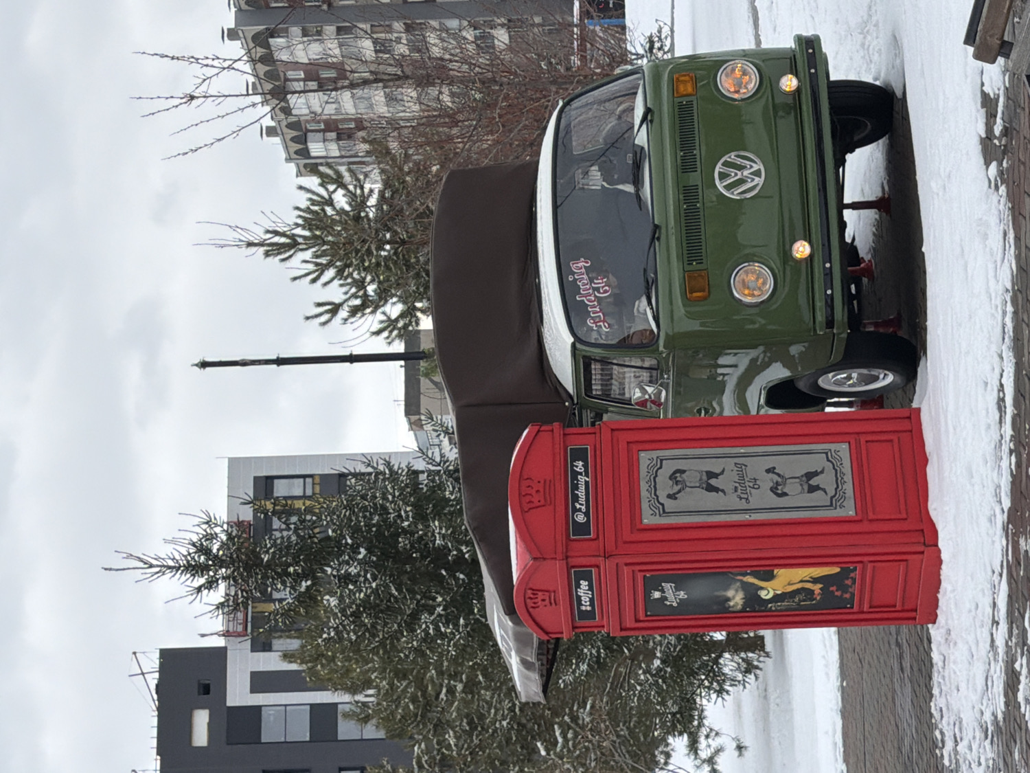 This outdoor image, likely captured in Krasnoyarsk, Russia, presents a winter scene featuring a vintage green and white Volkswagen van parked next to a distinctive red kiosk. Snow covers the ground and parts of the pavement, indicating cold weather, under an overcast sky during daytime.
The main subject is a classic dark green Volkswagen T2 or similar vintage bus, with a white roof. It features the iconic VW emblem on its front grille and round headlights with amber turn signals. The windshield displays a stylized sticker that reads "Ludwig 64". The van appears to be stationary, possibly acting as a mobile business or being set up for one. Red equipment, possibly jacks or supports, is visible beneath its front bumper.
Adjacent to the van is a bright red, British phone booth-style kiosk, which appears to be a coffee stand. It has several panels with text and graphics. One panel prominently features "@Ludwig.64" and "#coffee". Another larger panel displays "Ludwig 64" along with two silhouette figures, possibly boxers or strongmen, on a metallic-looking background. A lower panel includes "Ludwig 64" again, alongside an artistic illustration of a woman, perhaps in a classical style, implying a brand identity for the "Ludwig 64" establishment.
In the background, several multi-story buildings of varying architectural styles are visible, some with modern facades and others appearing older. Bare branches of deciduous trees and green evergreen trees, lightly dusted with snow, are scattered throughout the scene. A tall black pole, possibly a streetlight or crane, is also visible. There are no people present in the image.