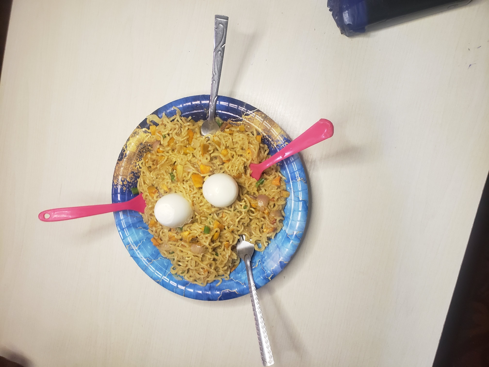 A close-up, overhead shot depicts a blue paper plate filled with instant noodles and two hard-boiled eggs. Three forks and two plastic spoons are scattered around the plate, some partially submerged in the noodles. The plate is positioned on a light-colored, textured surface. A dark object, possibly a phone, is partially visible in the lower right corner. The setting appears to be a domestic environment.