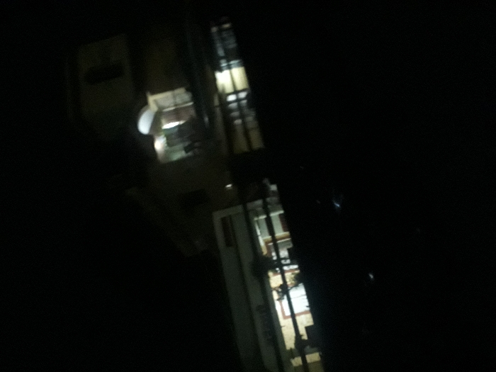 The image depicts a dimly lit scene taken at night from a low angle, likely from a moving vehicle, showcasing a residential building in Yavatmal, India, at the coordinates 20.38° N, 78.117° E. The building is partially visible, with a lit window on the upper floor and a second window on the lower floor partially illuminated. Several blurred figures can be seen standing near the front of the building, but their details are unclear. The environment is dark, with limited visibility, suggesting that it is a residential area with a building positioned on a street or road.