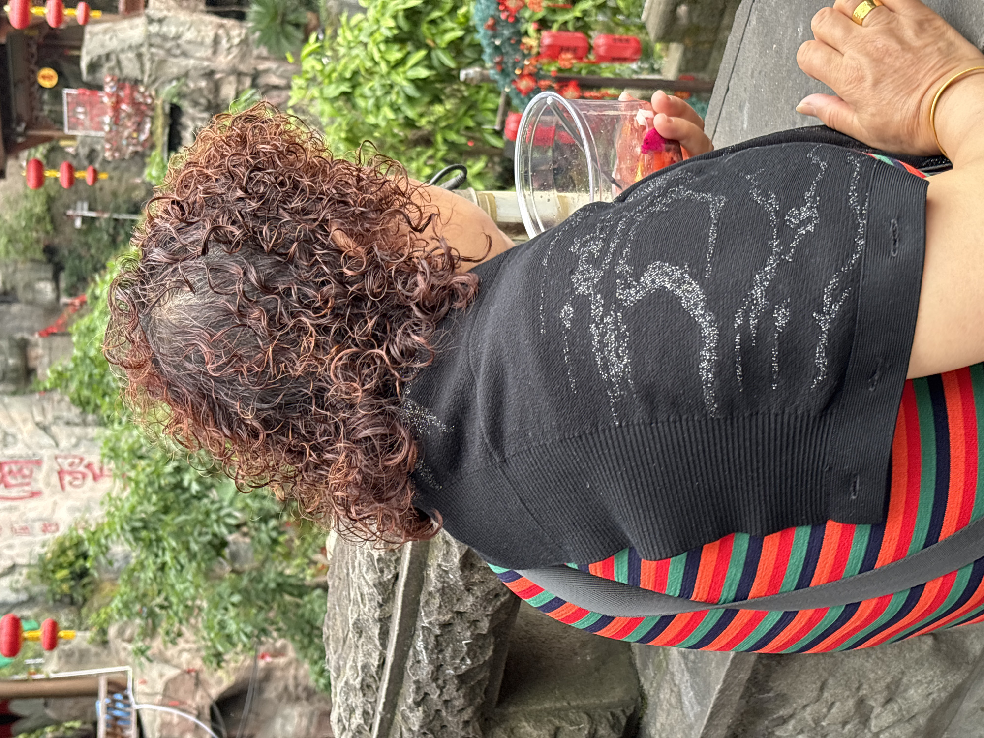 A slightly canted, mid-shot shows a person with curly reddish-brown hair sitting beside a rough, gray stone structure. The person is wearing a black, knitted top with silver glittery designs, and a striped dress or shirt with alternating bands of red, green, and dark blue. A gray strap crosses their back. They are holding a clear plastic cup containing a pink liquid. Their left hand rests on a gray surface. The hand has a gold ring on one finger and a gold bracelet on the wrist. The background consists of greenery, stone structures, and red lantern-like decorations. The location appears to be Yuzhong District, China.