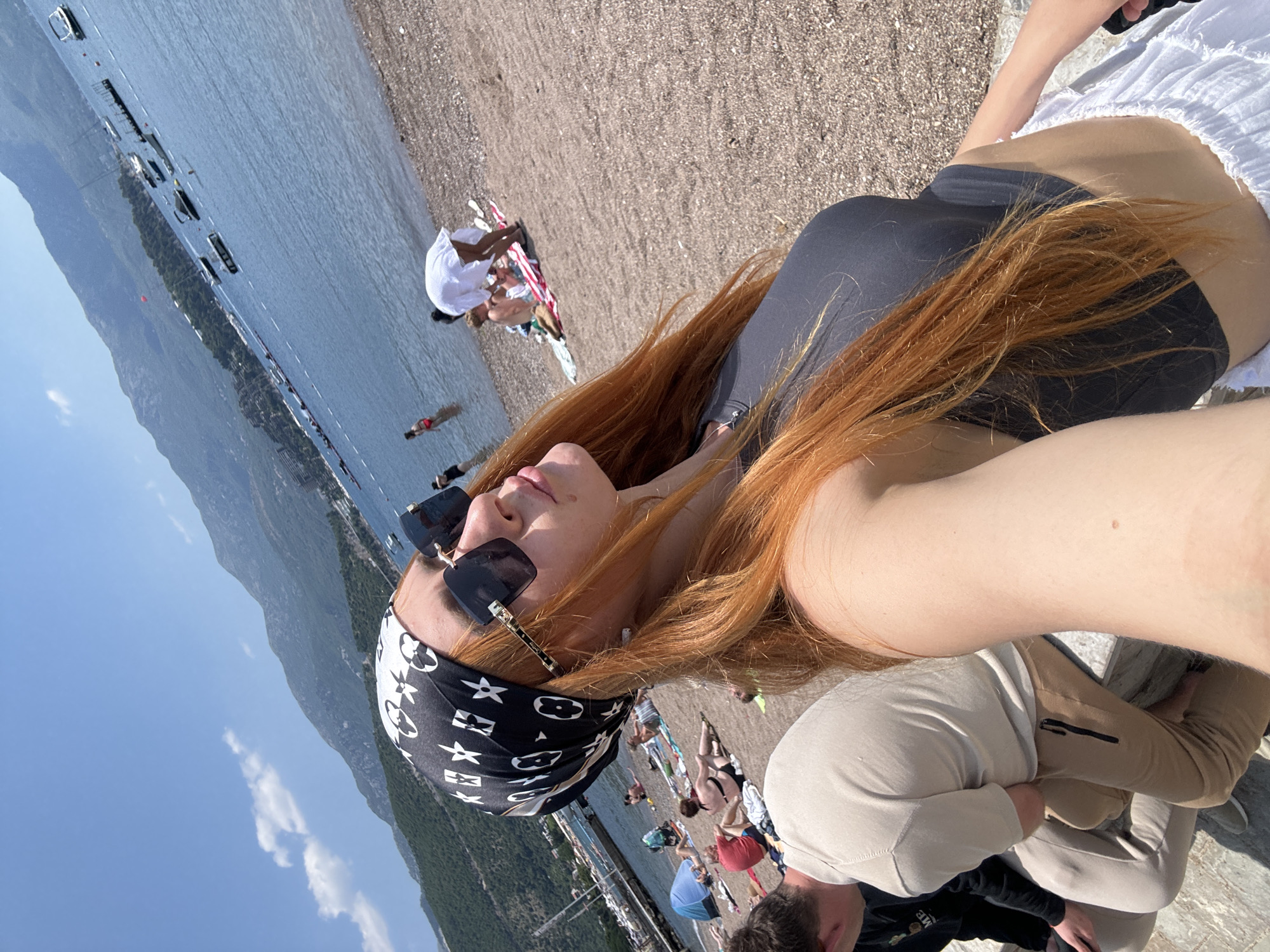 A woman with long, red hair and a black and white bandana is taking a selfie on a sunny day at a pebble beach in Budva, Montenegro. She is wearing large, square sunglasses and a dark grey tank top, with a white ruffled skirt visible at the bottom of the frame. In the background, the blue sea is dotted with several boats. The beach is populated with other people, some lying on towels, others wading in the water. In the distance, lush green mountains form the backdrop under a clear blue sky with a few scattered clouds. Two men are visible in the foreground to the left, one wearing a beige long-sleeved shirt and tan sweatpants, and the other wearing a black jacket.