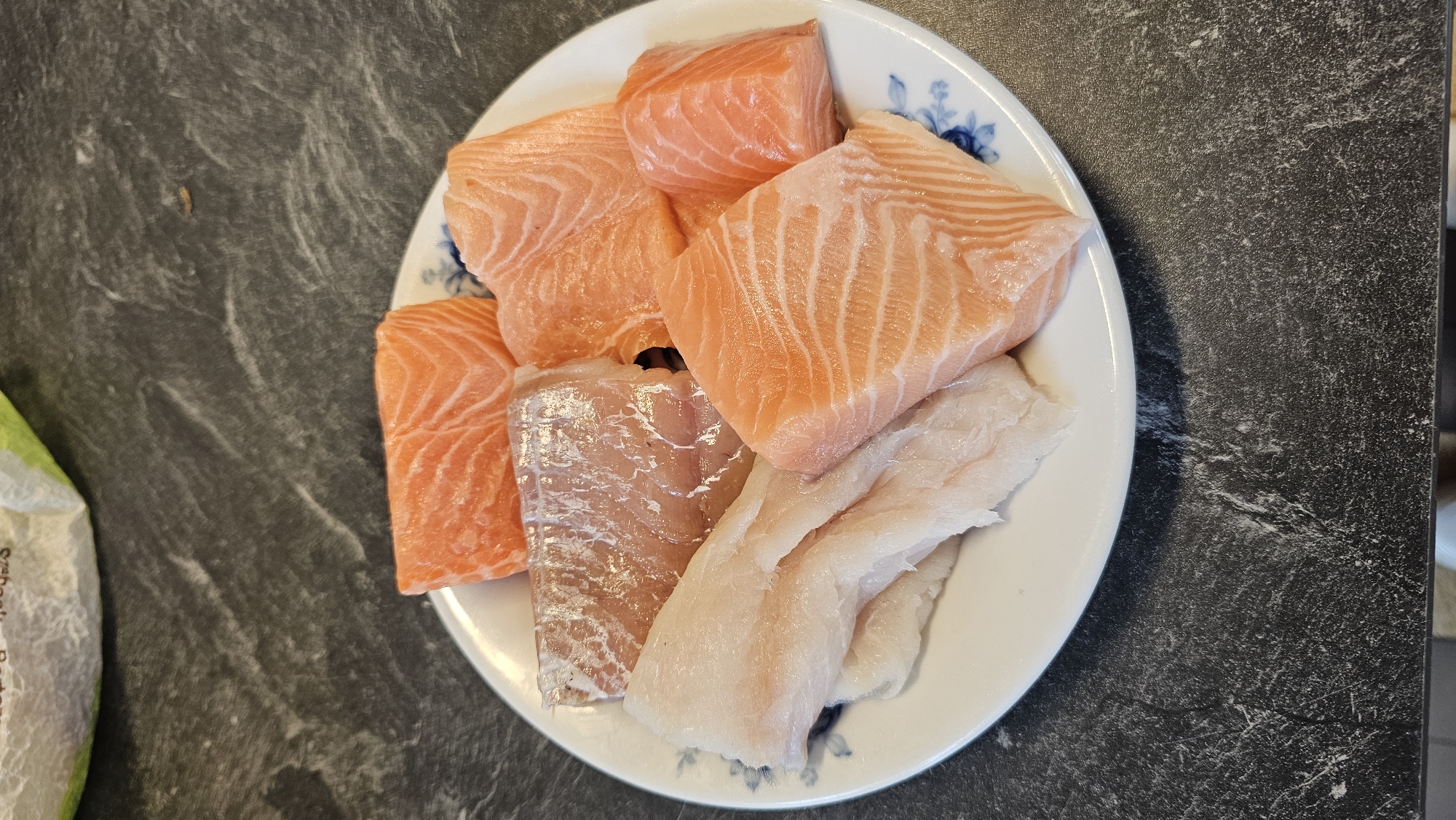 A top-down view shows a white, round plate filled with various raw fish fillets, resting on a dark, mottled countertop or table. The plate features a subtle blue floral pattern visible on its rim in several areas.
The majority of the plate is occupied by bright orange-pink salmon fillets, characterized by their distinct parallel fat lines. There are approximately five pieces of salmon, varying in size and shape, suggesting they have been cut from larger fillets. Alongside the salmon, there are at least two pieces of white fish fillets, which are paler in color and have a smoother, less textured appearance compared to the salmon. These white fish pieces are flatter and appear to be from a different type of fish, such as cod or similar.
The dark surface beneath the plate exhibits a textured, somewhat streaky pattern, resembling dark stone or a laminate material. On the far left of the image, a green and white partially visible bag or packaging can be seen, with some faint, indistinct text printed on it which is unreadable.
The arrangement suggests the fish are prepared and ready for cooking, possibly for a meal in an indoor kitchen setting, in Philippeville, Belgium. No other specific location or time details are discernible from the image.