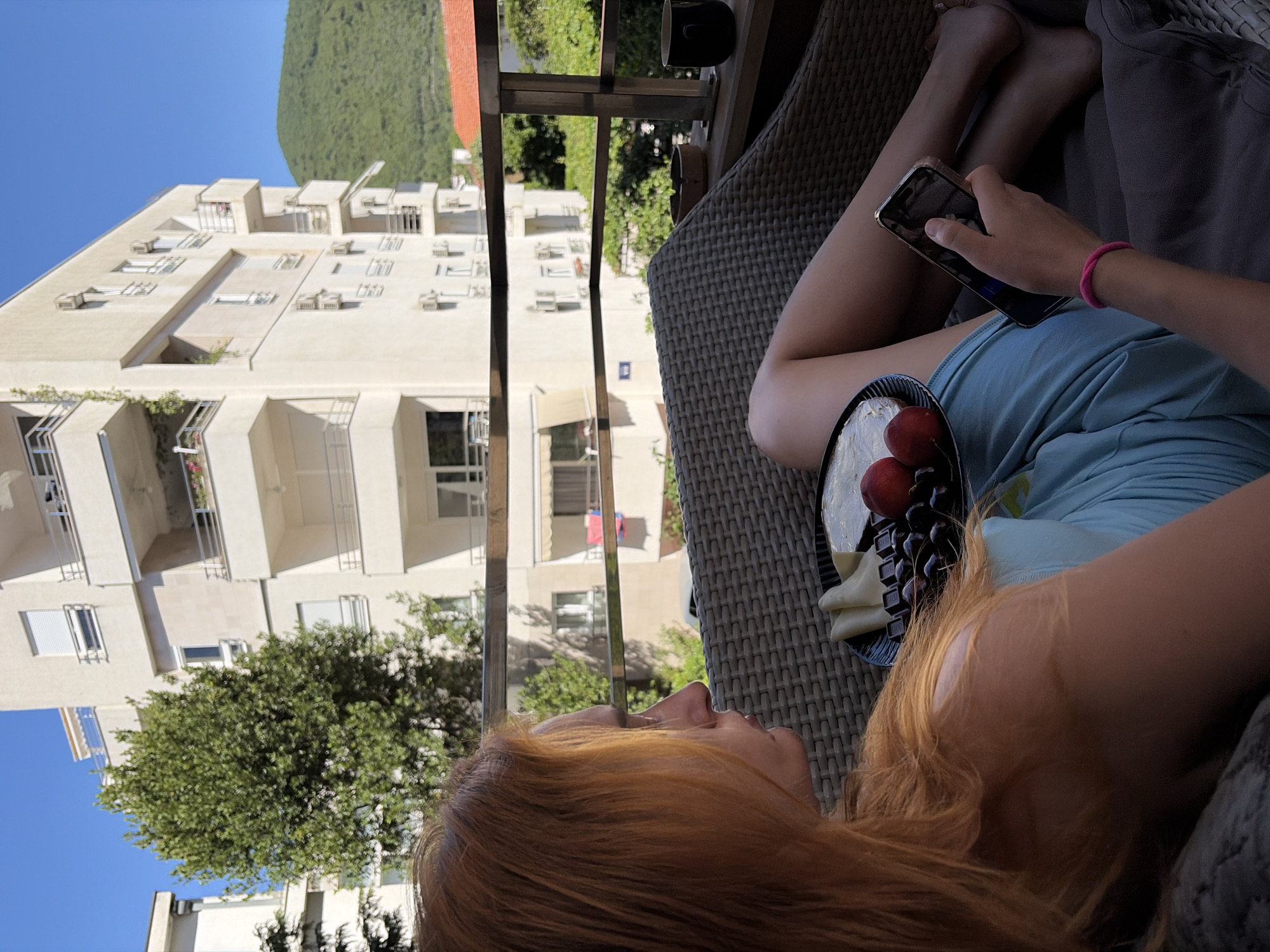 A young woman with long, auburn hair sits on a woven chair on a balcony in Budva, Montenegro. She is wearing a light blue sundress and has a pink bracelet on her wrist. In her lap, she holds a smartphone with a clear case and is looking at the screen. A plate of food, including cheese, cherries, and chocolate, rests on the chair beside her. In the background, a white apartment building with balconies and air conditioning units is visible, along with lush green trees and a mountain. The sky is clear and blue.