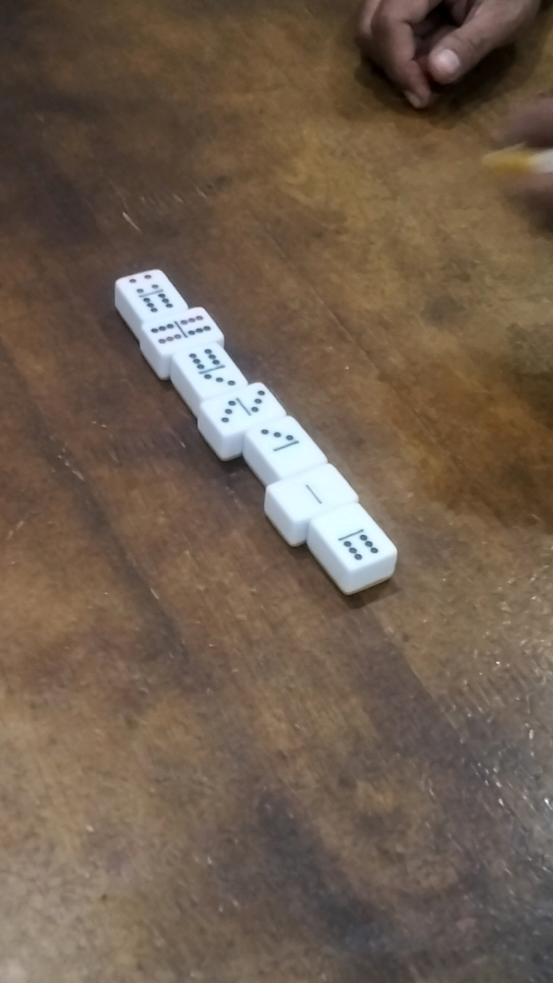 The image shows a close-up, angled view of several white dominoes laid out in a line on a wooden surface. The dominoes are positioned diagonally across the frame. The wooden surface appears to be a table, with visible grain and some scuff marks. In the upper right corner, parts of two human hands are visible. One hand appears to be reaching towards the dominoes, and the other is partially obscured. The lighting suggests an indoor setting. There are no clear indicators of the specific location within Lhokseumawe, Indonesia, beyond it being a general indoor environment where a game of dominoes is being played.