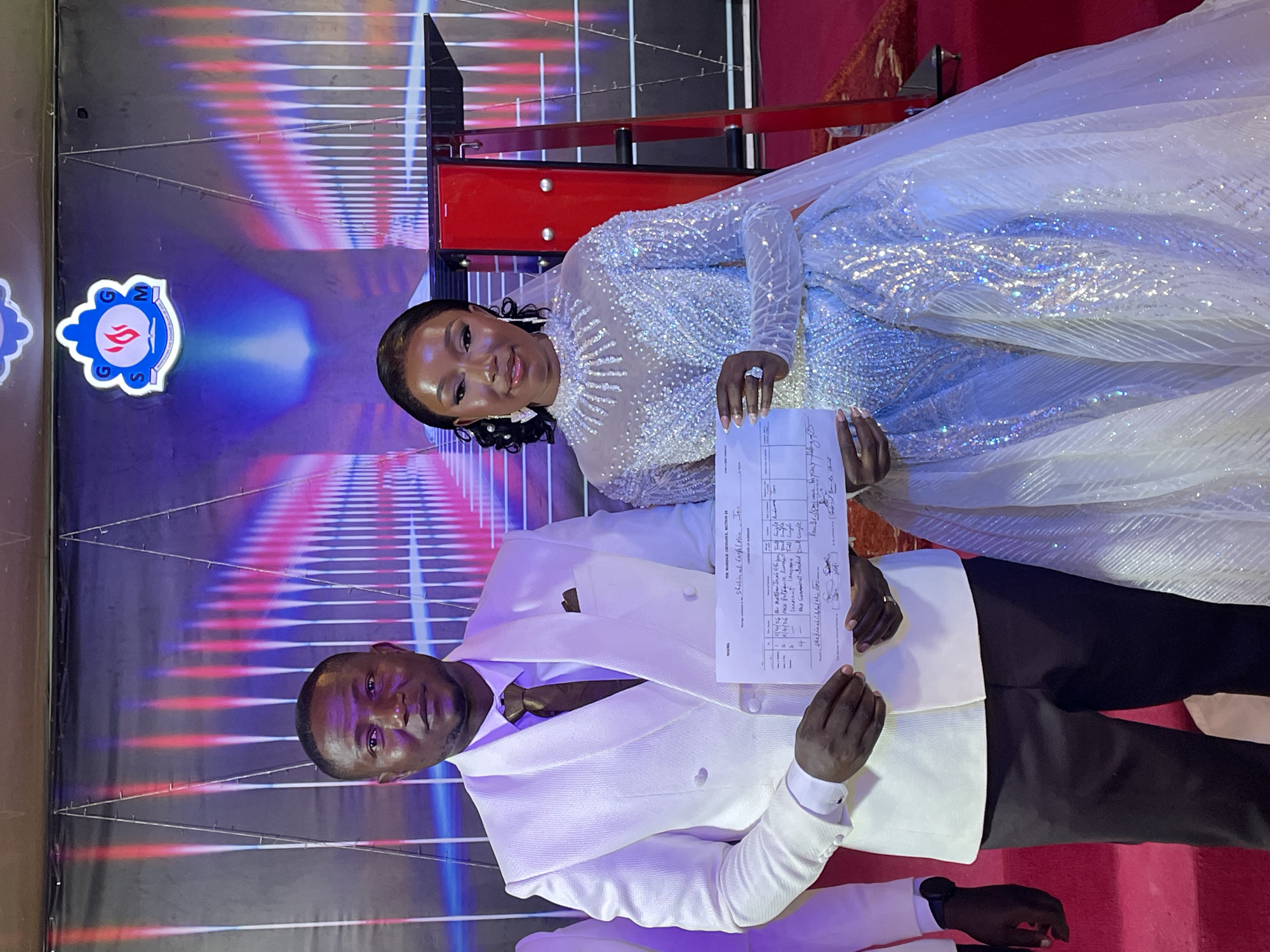 A man and a woman are standing in what appears to be a wedding venue in the city of Unknown, Unknown. The woman is wearing a sparkling white wedding gown and holding a document, possibly a marriage certificate, with the man. The man is wearing a white suit jacket, brown tie and black pants. The document reads:
Marriage CRESCe, Section IM
Shekinah Gospel Min. Jes
11/4/26 th Matthew Jones Eigen Full Song
4/4/26 Miss Patience Zurna Full
4- Innocent Jangara Fall single
the Guremical backed foul cingle
Shekinah
The background includes a stage with red and white design elements. There is a logo visible in the upper left corner of the image. A red carpet is also visible near the top right of the frame.