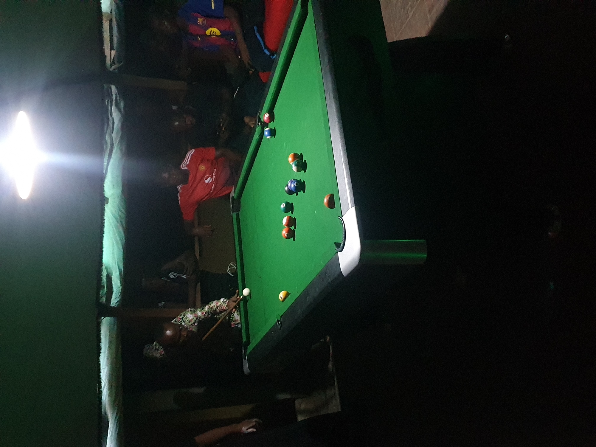 The image captures an indoor scene at what appears to be a recreational establishment in Lokoja, Nigeria, likely during the evening or night due to the dim, artificial lighting. The central focus is a green pool table where a game is in progress.
Several people are gathered around the table. One individual, wearing a patterned long-sleeved shirt, is actively taking a shot, holding a pool cue with both hands and leaning over the table. A white cue ball is positioned near the cue, and several colored pool balls, including solids and stripes, are scattered across the green felt. Among the balls, two red, two blue, two orange, one yellow, and one black ball are clearly visible.
Other people are partially visible in the background and around the table, observing the game. One person is wearing a red Manchester United football jersey with a white sponsor logo, where the letters "ODA" are partially visible. Another person in the background is wearing a blue and red FC Barcelona jersey with a yellow-gold crest or logo on the chest. A hand, possibly chalking a cue, is visible in the lower left corner.
A very bright, rectangular light source is prominent on the left side of the frame, casting a strong glare and illuminating parts of the scene, while other areas remain quite dark. The overall atmosphere suggests a casual and engaging social activity.