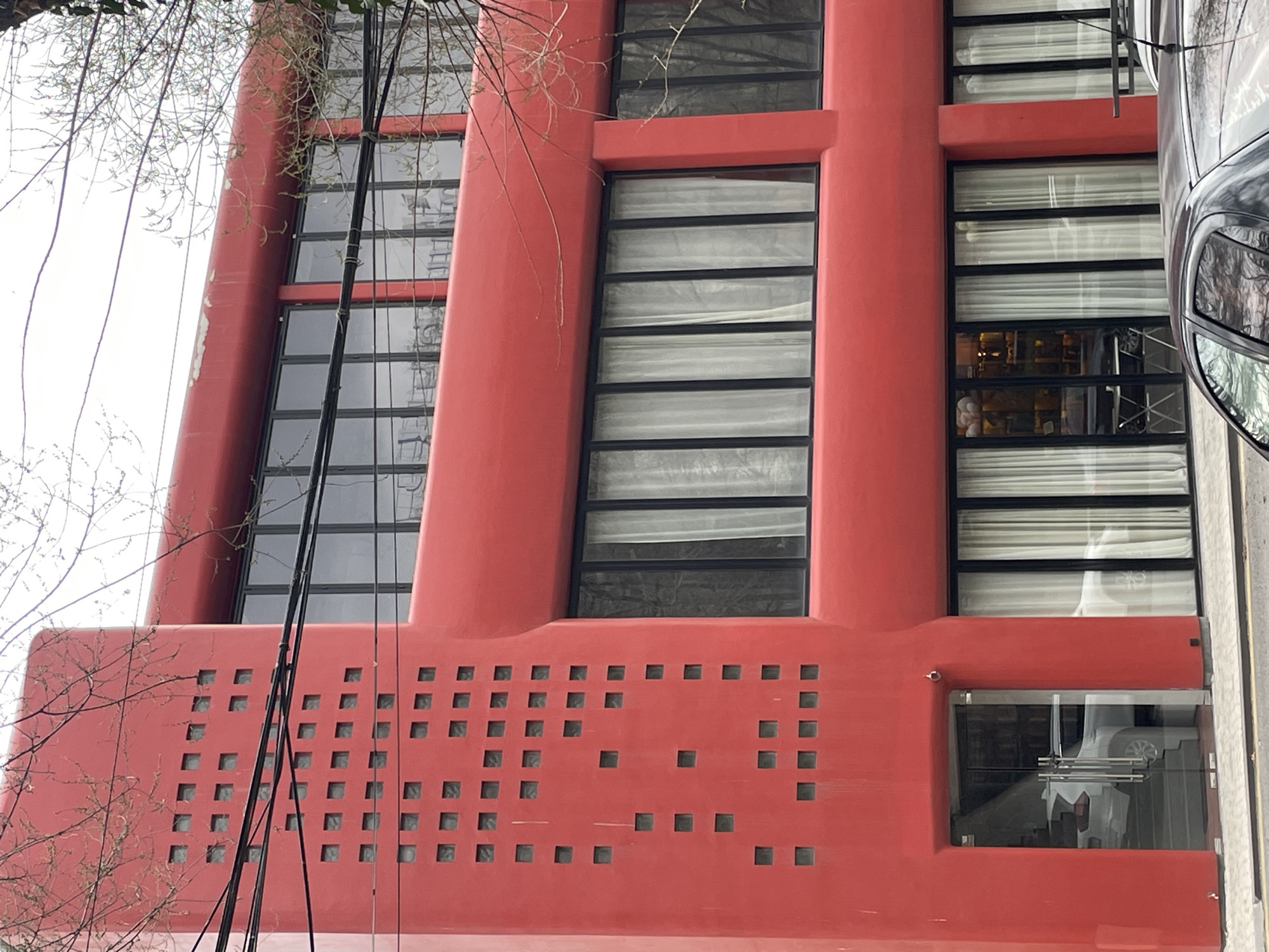 The image captures a close-up, low-angle view of a distinctive red building facade, likely in Yerevan, Armenia. The building features a modern design with a series of vertically stacked windows, framed by robust red columns. The windows have horizontal black bars, partially obscured by white curtains or blinds inside.
The base of the building is adorned with a patterned array of square openings, adding a decorative element to the facade. A single window near the base appears to show the interior of the building.
To the right of the building, a black car is parked, its reflective surface showing the surrounding environment. Bare tree branches are visible on the left side of the frame, indicating the season may be late autumn or winter.
Black cables run diagonally across the building's surface, adding a visual disruption. The building's red color is the dominant feature, contrasting with the gray sky and the muted tones of the surrounding environment.