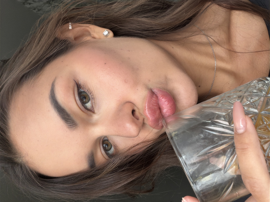 A woman holds a glass, tilted at an angle, to her lips. She has long brown hair and fair skin. She is wearing earrings. She has dark eyebrows and her eyes are hazel in color. The glass is clear and seems to contain an amber liquid. She wears a delicate silver chain around her neck. One hand is visible holding the glass, with a long, manicured nail. The background is blurry, but seems to be dark gray.