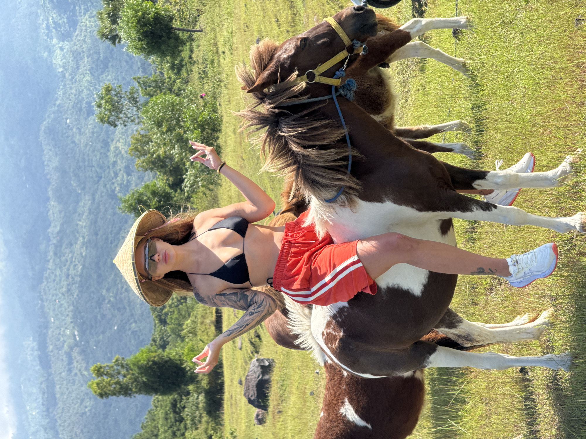A woman is straddling a horse in an outdoor setting. The location appears to be a grassy field in Kubu, Indonesia, with mountains and trees visible in the background. The woman is wearing a black bikini top and red shorts with white stripes, a straw hat, and sunglasses. She has tattoos on her arm. The horse is brown and white. There is another horse in the background.
