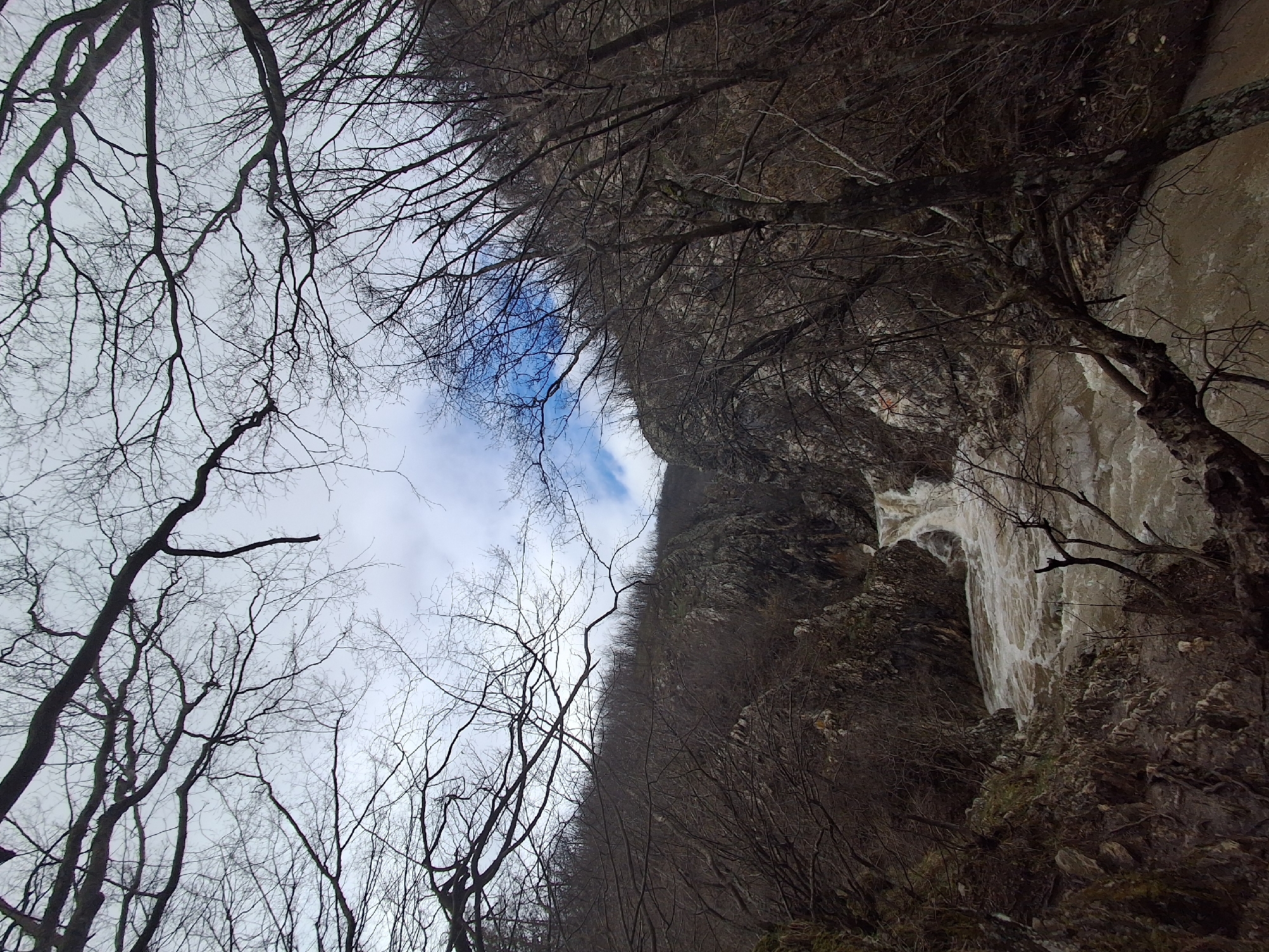 The image shows a waterfall and river flowing over a cliffside. The viewer is looking up toward the top of the waterfall with bare trees visible on both sides of the frame. The sky is visible through the trees and is mostly overcast with some blue sky showing. The photograph was taken in Medven, Bulgaria.