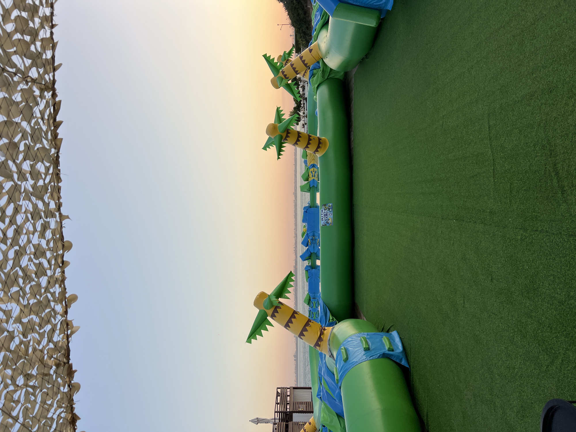 In Fontanka, Ukraine, a vibrant, inflatable water park is set up on a patch of artificial grass, facing the sea. The water park features large, green inflatable tubes connected to form an obstacle course, with stylized palm tree inflatables adorning its perimeter. A camouflage net stretches across the top of the frame, providing shade. In the background, the calm sea meets a pale orange and blue sky as the sun begins to set. A few buildings and structures are visible along the coastline, suggesting a developed resort or recreational area. The scene is devoid of any people, presenting a tranquil, pre-activity snapshot of the outdoor attraction.