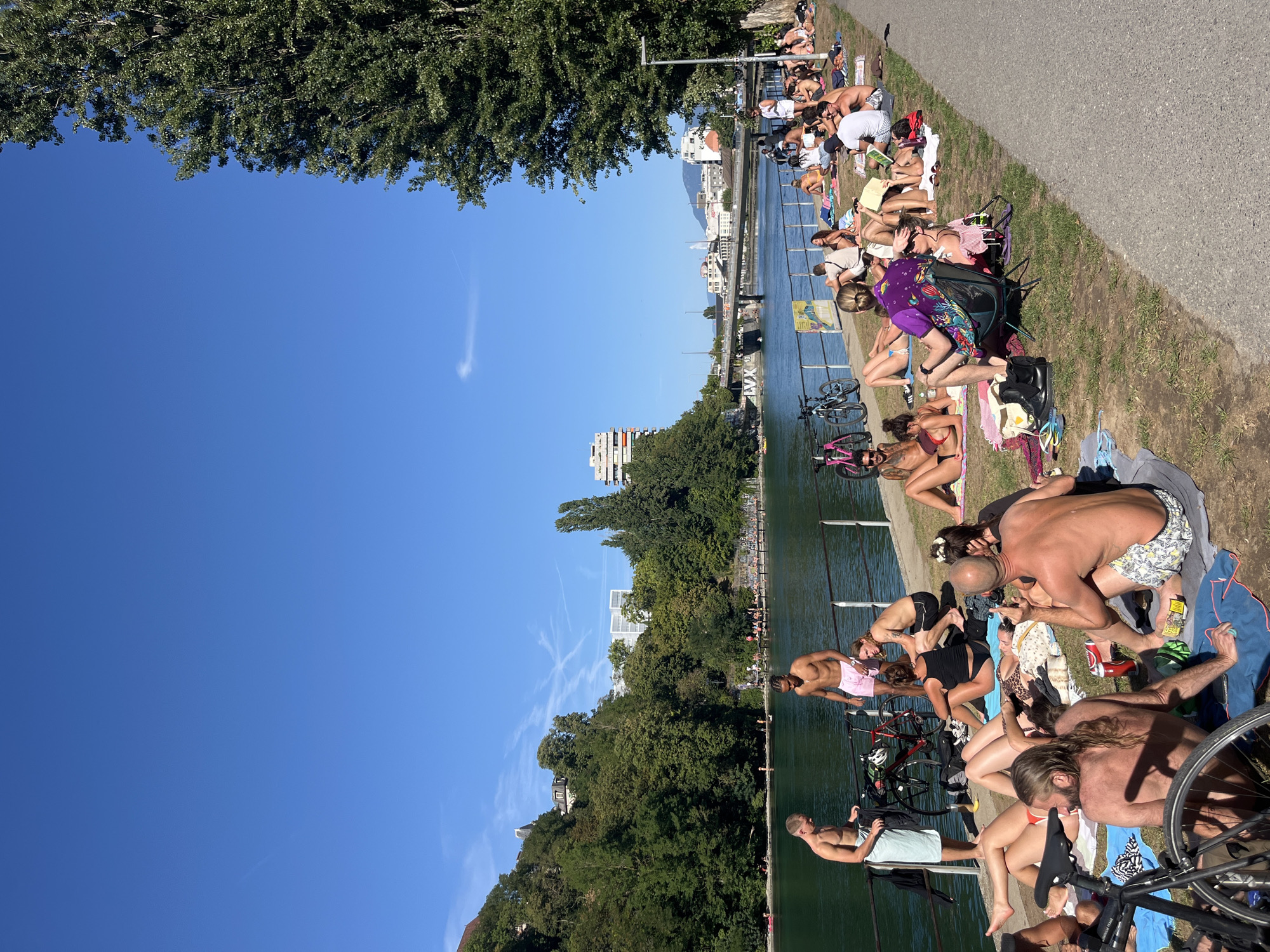 The image depicts a sunny day scene along the Rhone River in Geneva, Switzerland, near the Jet d'Eau. A diverse group of people are sunbathing and relaxing on the grassy bank. Several individuals are wearing swimwear, with many lying on towels or blankets. Some are reading, others are engaged in conversation, and a few are actively sunbathing. Bicycles are parked near the riverbank, suggesting recreational activity. In the background, the river is visible, with a bridge spanning across it, and urban buildings are seen on the opposite shore. The sky is clear and blue, indicative of good weather. The overall atmosphere is one of leisure and enjoyment of the outdoors in an urban setting.