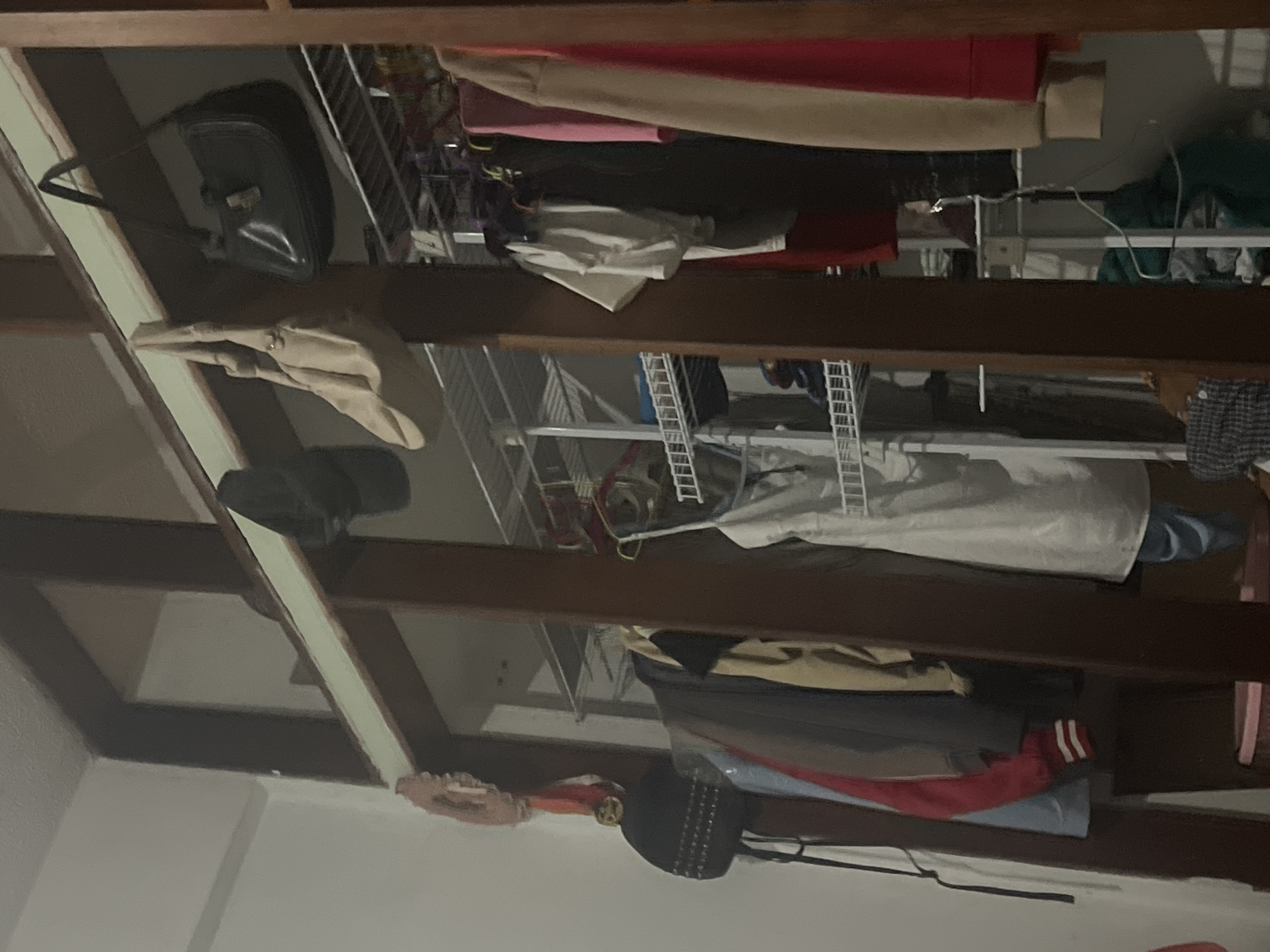 The image depicts an interior view of a closet or wardrobe area, likely in a residential setting. The scene is dimly lit and cluttered with clothing and accessories. In the upper section, several bags are visible hanging from a wooden structure. On the left, a black studded handbag is hanging next to a decorative circular item. To its right, a light-colored, possibly beige, handbag is hanging, followed by a black leather handbag with a flap closure. Below these bags, clothing items are arranged on hangers within the closet. These include various garments such as jackets, sweaters, and dresses, with a white dress prominently displayed in the center. Shelving units are visible behind the hanging clothes, containing folded items and possibly more bags. The overall impression is one of a functional but somewhat disorganized storage space. The location is specified as Santa Ana, Venezuela.