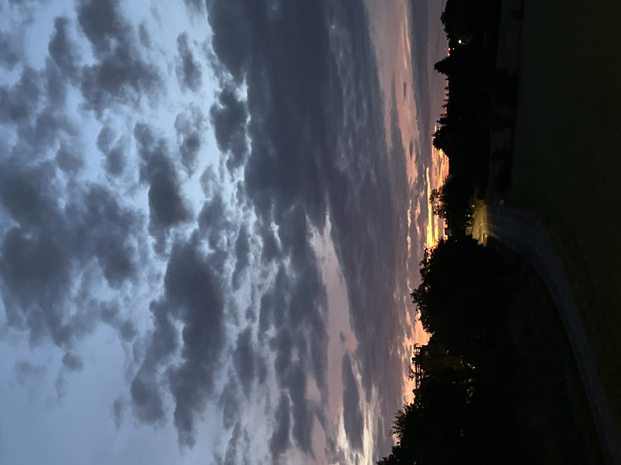 The image shows a twilight sky over a rural landscape, possibly in the area of Leudelange, Luxembourg. The sky is filled with dramatic, dark clouds, with streaks of orange and pink light indicating the setting or rising sun. Silhouetted trees and a faint outline of buildings are visible against the horizon. A road curves through the landscape, with a few distant lights suggesting human habitation. The foreground is dark and appears to be grassy fields or parkland. There are no people or animals visible in the image.