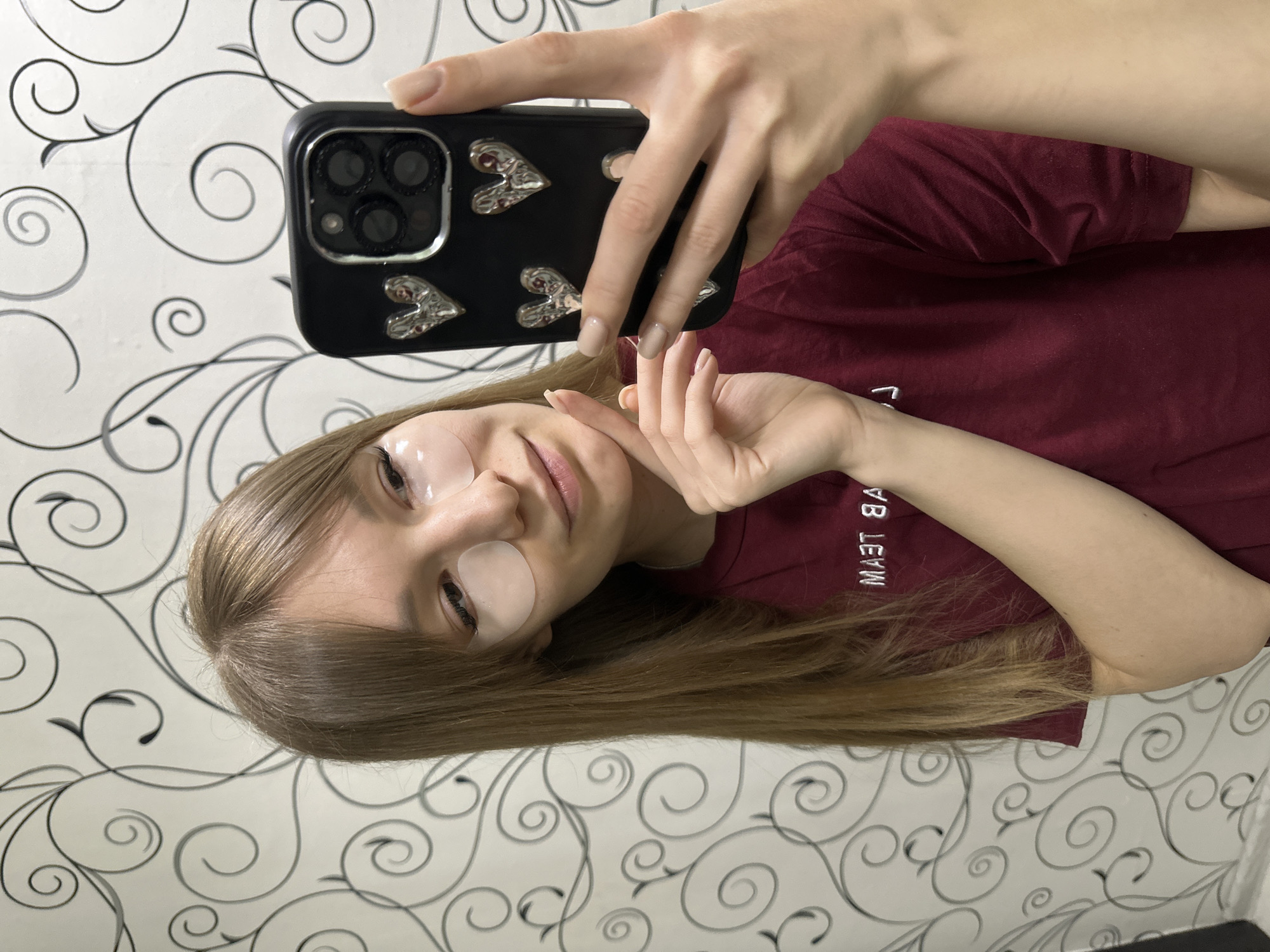 A young woman is taking a selfie indoors. She is wearing eye patches and a maroon shirt. She holds a black smartphone with a heart-decorated case up to take the picture. The background is a white wall with a gray and black swirling vine pattern. The scene is located in Chaikovsky, Russia.