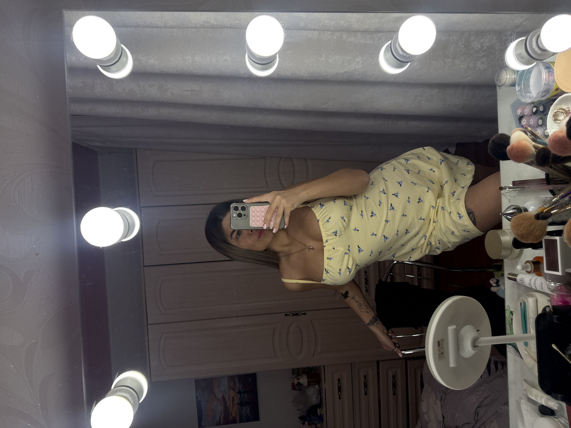 A woman in a yellow dress with blue bird patterns takes a selfie in a mirror with Hollywood-style lighting. The mirror is positioned above a makeup table cluttered with cosmetics and brushes. The reflection reveals a bedroom setting with white cabinetry and a painting of trees and a sunset, which places the scene indoors. This photo seems to be taken in a residential space, possibly in Chaikovsky, Russia.