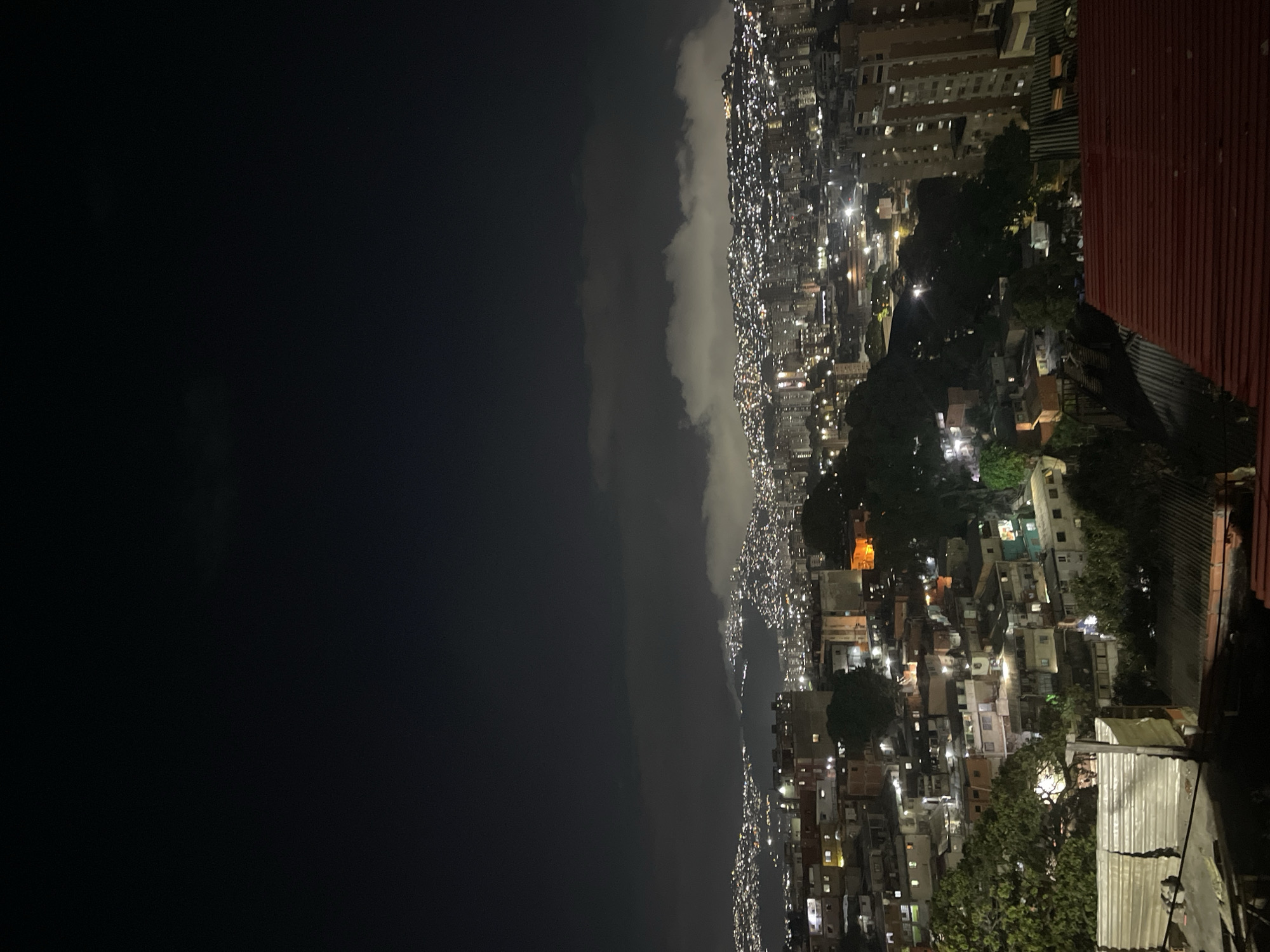 The image shows a night view of Caracas, Venezuela, from an elevated position, likely a rooftop. The city is densely populated with buildings of varying heights, including high-rise apartments and smaller houses, illuminated by streetlights and lights emanating from the buildings. A dark sky fills most of the left side of the frame, while a layer of clouds stretches along the horizon where the city ends. The scene includes residential areas and possibly commercial districts, indicated by the density of lights. In the foreground, there is a corrugated metal roof, likely of the building where the photo was taken. The city's terrain appears hilly, as suggested by the elevation changes. The image does not show any people.