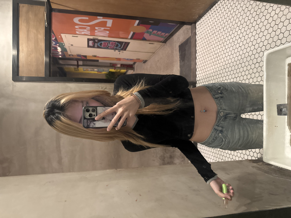 An individual, presumed female, with long, two-tone hair (blonde with dark roots), is depicted in a mirror selfie. The individual wears a black long-sleeved crop top exposing the midriff, light blue low-rise jeans, and exhibits a belly button piercing featuring a light blue gem. A dark-colored smartphone, held in the individual's right hand (as seen in the mirror), partially obscures the face. The phone case displays a patterned design around its multi-lens camera module. The individual's left hand (as seen in the mirror) holds a green-handled brush-like object, pointed towards a textured, light beige wall. The floor features white hexagonal mosaic tiles with dark grout. The lower foreground includes the partial view of a white sink or counter, containing dark residue. Beyond the individual's reflection, a doorway with a dark frame opens into what appears to be a brightly lit corridor. This corridor's walls are adorned with large, vertically oriented posters, predominantly orange and yellow, displaying reversed text and graphics, including "CAR" and "S PON". A dark mat is visible on the corridor floor just past the doorway. The scene is an interior setting within the city of Liangjiang, China.