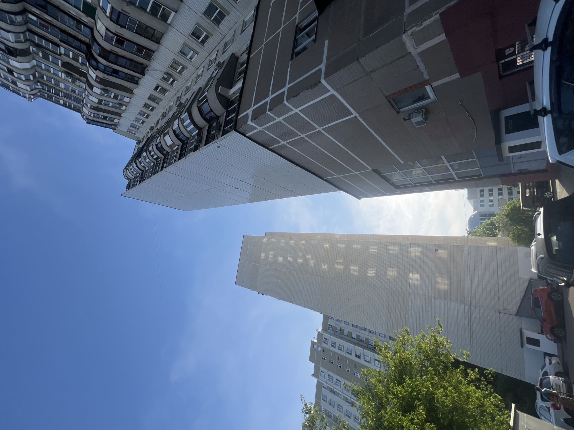 The image shows a view of buildings in what appears to be a densely populated urban environment. The buildings are predominantly high-rise apartment blocks with a variety of architectural styles. Some have curved balconies, while others are more angular and tiled. The sky is visible between the buildings, indicating a clear day. The photo also shows some street-level elements, including trees, parked cars, and possibly a sidewalk or road. Based on the architectural styles and urban density, it is highly probable that the location is in the city of Moscow, Russia.