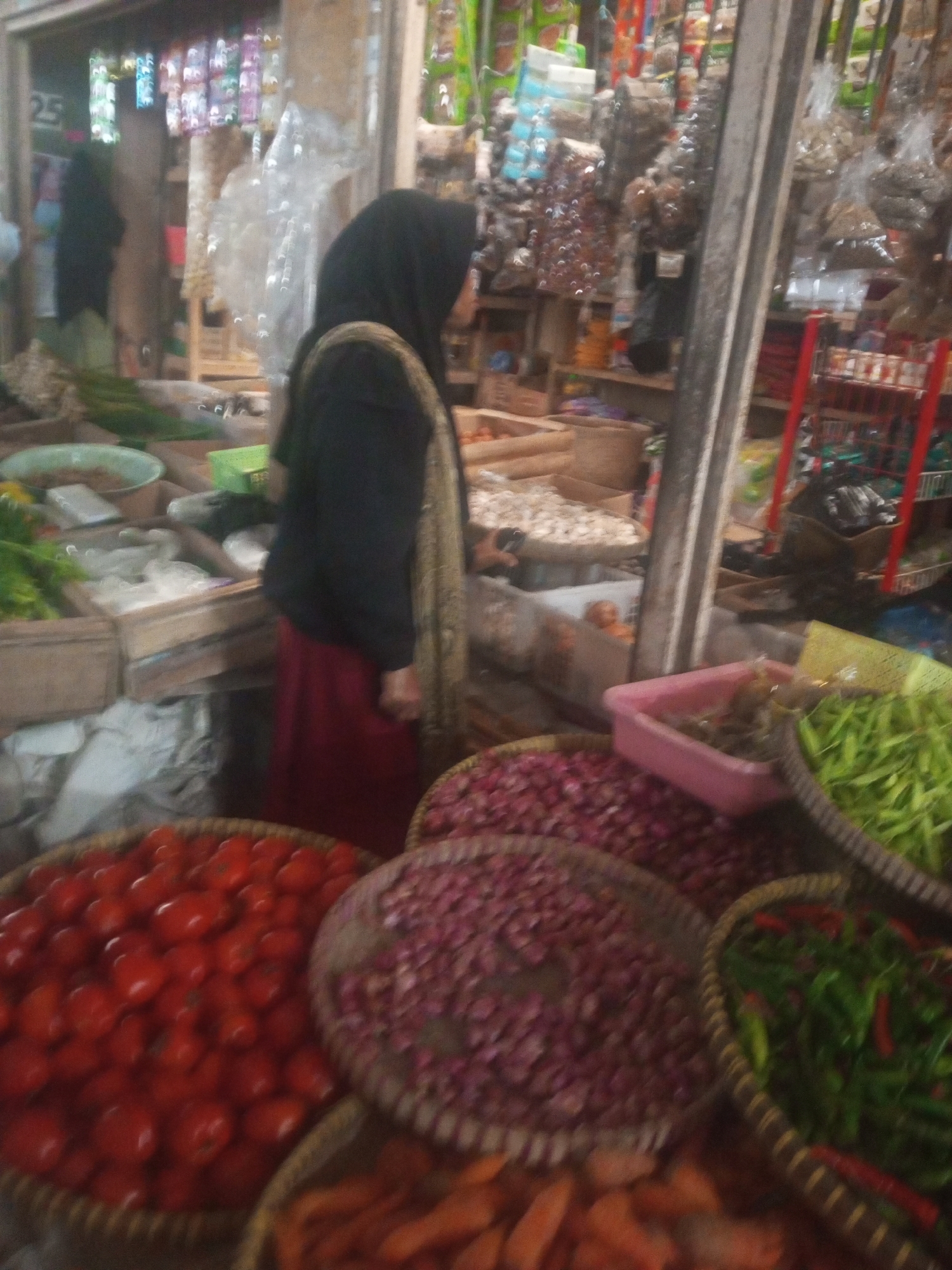 In the city of Kalibeber, Indonesia, a bustling market scene unfolds. In the foreground, a woman wearing a black hijab and a maroon skirt stands behind a display of fresh produce. Her right arm is bent, and her hand is near her side. She is positioned in front of a large woven basket filled with vibrant red tomatoes. To her left, another woven basket contains a mound of purple onions. To the right of the onions, a pink plastic container holds some green beans or similar produce, and further to the right, a basket overflows with green chili peppers and some red ones.
The background reveals a vibrant market stall with shelves laden with various goods, including packaged items and dried goods hanging from above. The market appears to be covered, with wooden structures and shelves visible, suggesting an indoor or semi-covered market setting. Sunlight or artificial light casts a bright illumination on some of the items, creating reflections and highlights. The overall impression is one of a lively marketplace offering a wide array of fresh produce and other commodities.