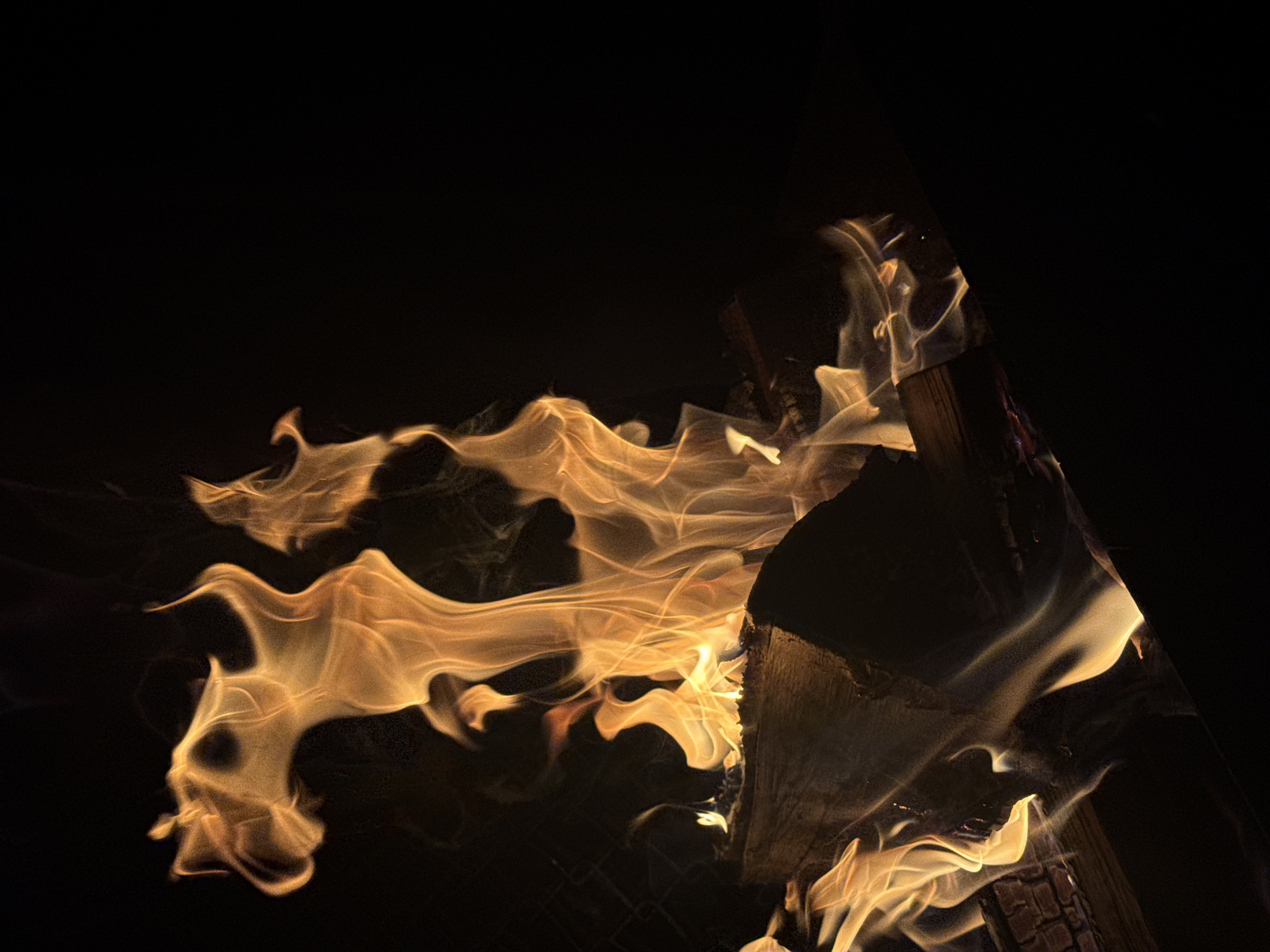 The image shows a close-up view of a campfire at night. The flames are a vibrant orange and yellow, with wisps of smoke rising into the dark sky. In the foreground, several pieces of wood are visible, some of which are burning. The overall impression is one of warmth and intensity, set against the backdrop of a dark, quiet night. The location is not specified in the image, and therefore cannot be determined.