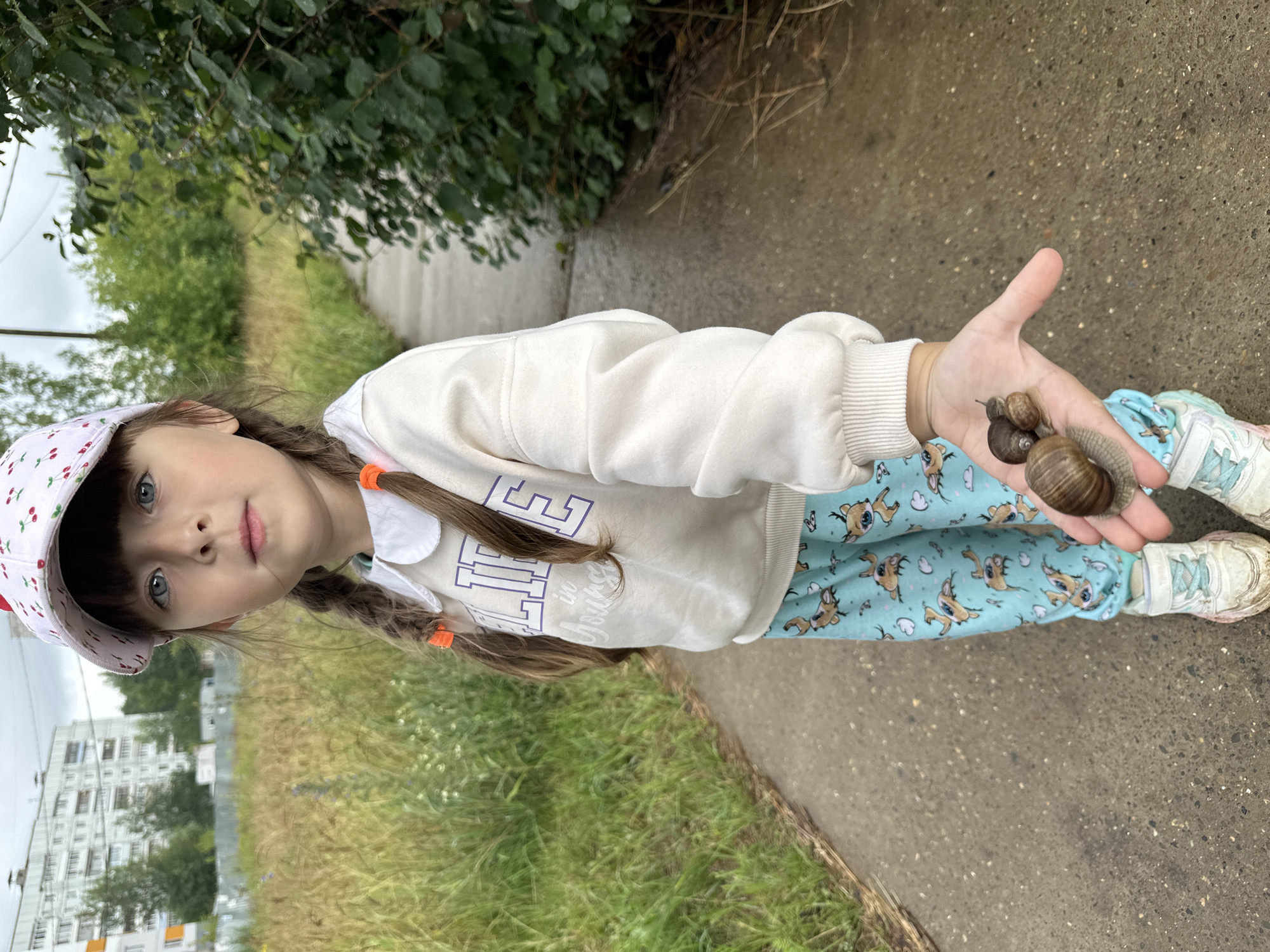 A young girl, appearing to be about five years old, stands on a paved path outdoors. She wears a white baseball cap with a cherry pattern, a cream-colored sweatshirt with the word "LIFE" and "in Young" printed on it, and light blue pants adorned with cartoon deer. Her hair is styled in two braids, each secured with an orange hair tie. In her right hand, held open, are three snails. The background shows a grassy field with some wildflowers, a dense green bush on the right side, and a multi-story building in the distance, characteristic of urban housing in Chaikovsky, Russia. The sky is overcast. The girl is looking directly at the camera with a neutral expression.