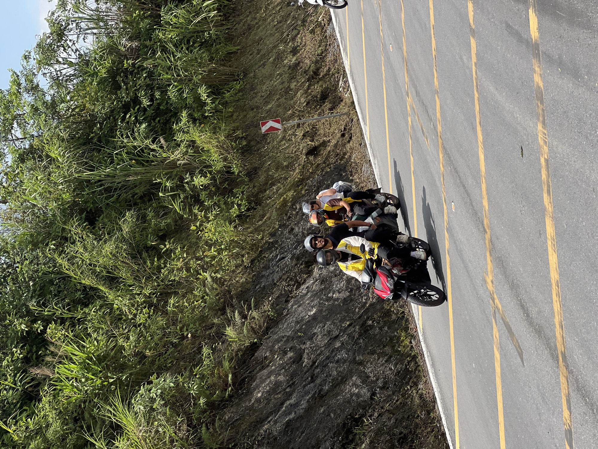 A group of four individuals, each wearing a helmet and riding on a red and black motorcycle, is traveling on a paved road in Quản Bạ Commune, Vietnam. The road has yellow lane markings. The motorcycles are traveling uphill along a winding road, with a steep, rocky incline covered in lush green vegetation on the left side and more dense foliage on the right. A directional sign indicating a sharp right curve is visible on the right side of the road. The sky above is clear and blue.
