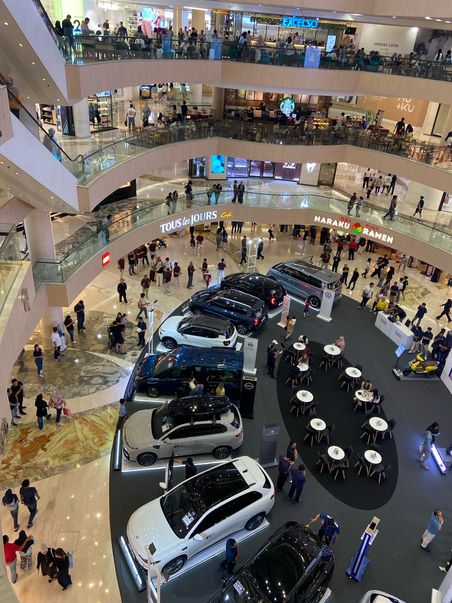 This is a high-angle, full-shot photograph taken inside a multi-level shopping mall, likely located in Surabaya, Indonesia, given the presence of the "EXCELSO" coffee shop, a popular Indonesian chain. The mall features several escalators and walkways, with various shops and dining establishments visible on different levels.
In the foreground, a car exhibition is set up on the ground floor. Several cars are on display, including white SUVs and a dark blue sedan. People are browsing the cars and interacting with what appear to be sales representatives. Some tables and chairs are also set up in this area, indicating a lounge or seating area.
Further back, the mall's interior is bustling with activity. People are seen walking on escalators, browsing shop windows, and dining at cafes. Notable signs include "TOUS les JOURS," a bakery chain, and "HARAKU RAMEN," a Japanese restaurant. On a higher level, a "Starbucks" is visible. The overall atmosphere is bright and busy, characteristic of a large commercial space. The architecture is modern, with polished floors and glass railings on the walkways.
