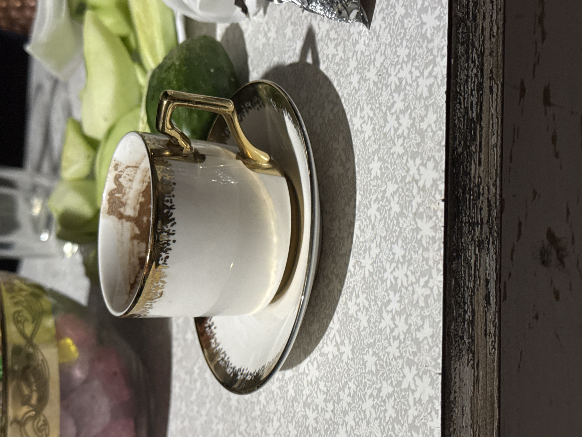 An overhead shot captures a close-up of a small, white ceramic coffee cup with a gold rim and handle, filled with a dark beverage, resting on a matching saucer. The cup and saucer are placed on a light gray textured surface, possibly a tablecloth, with a subtle floral pattern. In the background, out of focus, are several light green cucumber slices and what appears to be a round, dark green cucumber or similar vegetable. To the left, a blurred bowl with a gold rim contains pink-hued items, possibly fruits or candies. A glass is visible behind the bowl. The overall scene suggests a casual setting, possibly a meal or snack. The location is not discernible from the image itself.