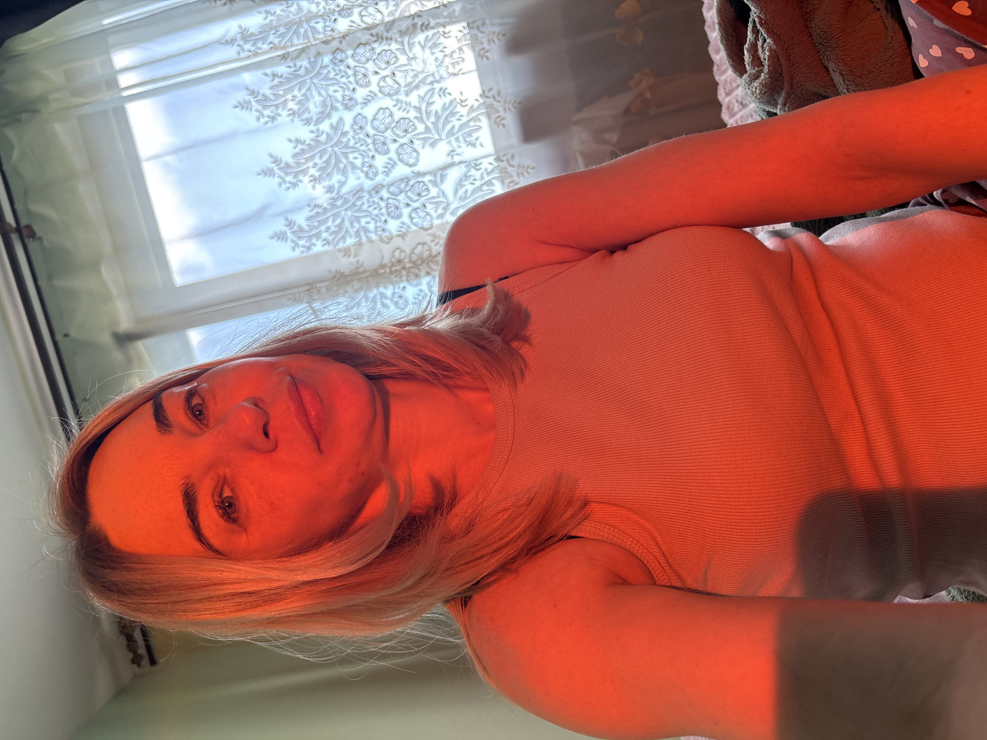 The image features a woman posing indoors, captured in a selfie-style shot. She is positioned with her upper body visible, including her face, arms, and torso. Her blonde hair is parted on the side and frames her face. She is wearing a light-colored, ribbed tank top or camisole.
The most prominent detail is the strong, warm orange-red light that illuminates the woman, giving her skin and clothing a vibrant reddish hue. This suggests the golden hour, likely sunset, with light streaming through a window. The background features a window with white, semi-sheer curtains adorned with an intricate floral and leaf pattern. Through the window, bright, diffuse daylight or a cloudy sky is visible.
The woman is looking directly at the camera with a neutral to slightly relaxed expression. Her eyes are open and focused. The image is oriented sideways, suggesting the photo was taken with the camera held horizontally or the subject was lying down. No specific activity beyond posing for the camera is evident. In the upper right corner, some dark fabric, possibly a blanket or clothing, is visible. There is no visible text in the image.