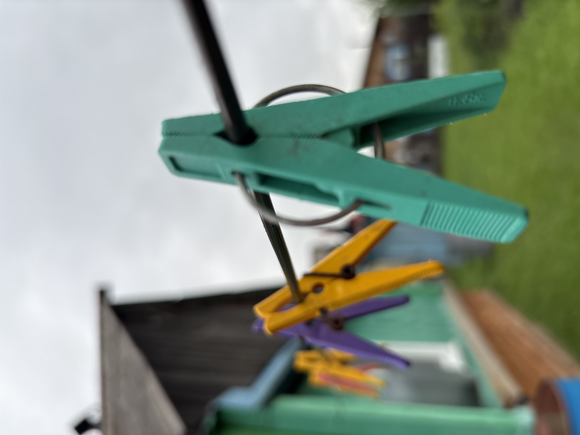 A close-up, shallow depth of field shot focuses on a green plastic clothespin clipped to a clothesline, which is stretched horizontally across the frame. The clothespin is in the foreground and is in sharp focus, showing a ribbed texture on its lower arms. Behind it, slightly out of focus, are several other clothespins of different colors—purple and yellow—also attached to the clothesline.
In the background, a weathered wooden house with a dark roof is visible on the left, partially obscured by the clothespins. The house has a green painted section on its lower visible part. To the right and further in the background, a blurred green lawn and some out-of-focus structures or vegetation are present. The sky above is overcast and light gray. The overall impression is of an outdoor setting, likely a backyard or laundry area. The image does not provide any explicit information to indicate it is specifically located in the city of Gonja, Russia.
