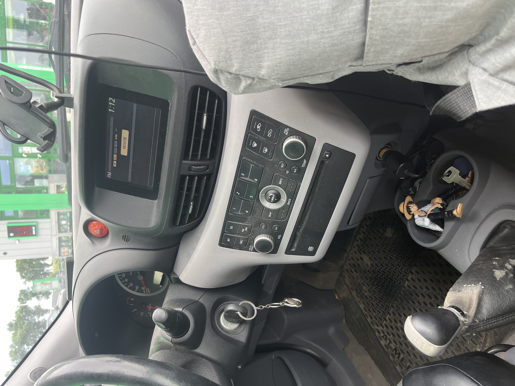 The image shows the interior of a car, likely a Nissan Almera based on the floor mat branding. The dashboard features a central infotainment system displaying "NOVCE" on its screen and a clock showing 1:12. To the left, a steering wheel and part of the instrument cluster are visible, with the odometer reading approximately 45,000 km. A hazard light button is illuminated. The gear shift lever is positioned in neutral. In the center console, a small figurine of Monkey D. Luffy from One Piece is seated in a cup holder, accompanied by keys. The driver's seat is partially visible, covered with a light grey fabric. The car appears to be parked outdoors, with a building and trees visible through the windshield, suggesting a location in Шчомысліцкі сельскі Савет, Belarus. The overall scene is of a parked vehicle with personal items inside.