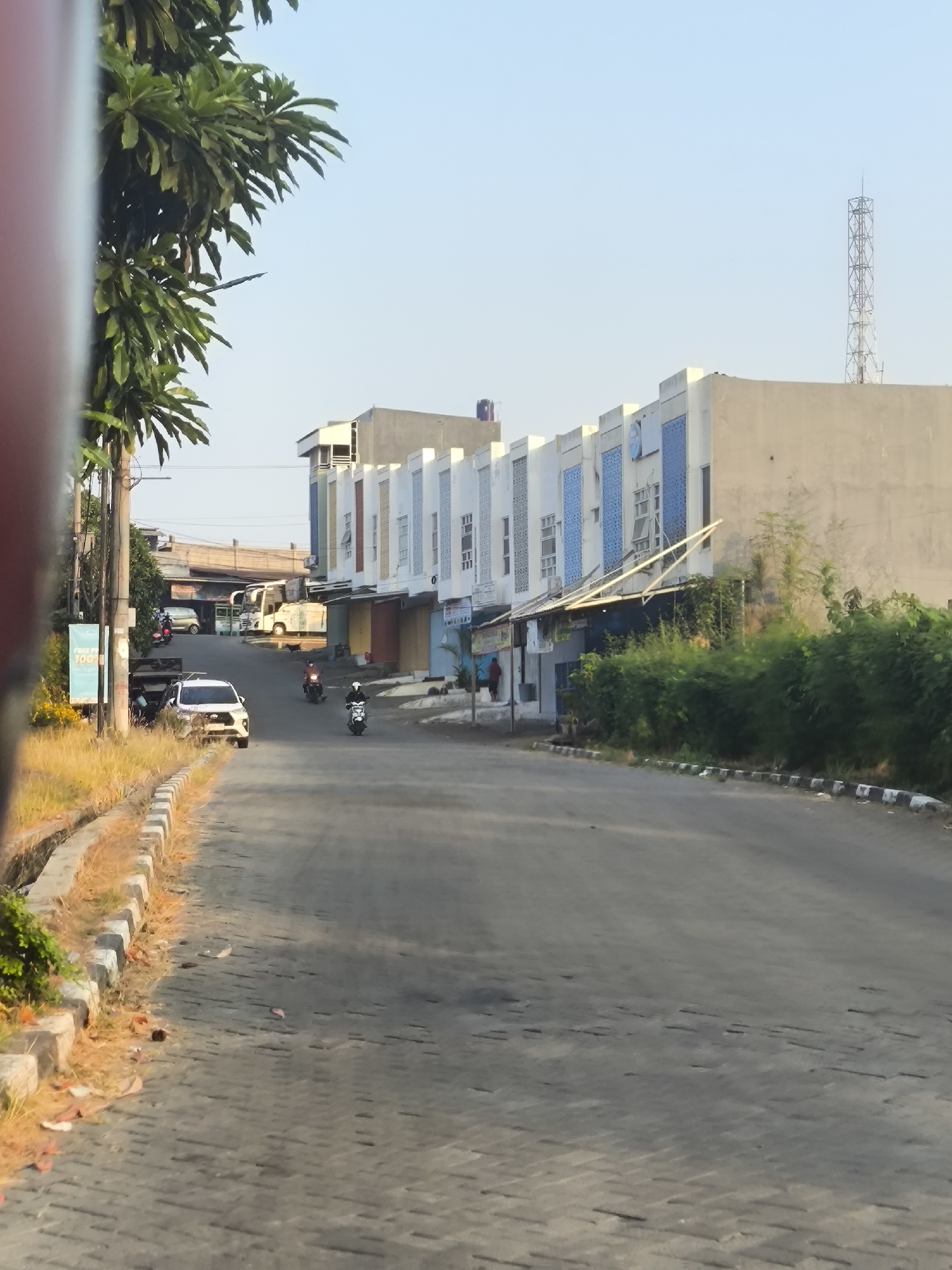 The image depicts a street scene in Cilegon, Indonesia, characterized by a row of commercial buildings and a clear sky. The road surface is paved with interlocking blocks, with a curb and a drainage ditch along the left side. Lush green trees and vegetation flank the roadside. Several vehicles, including cars and motorcycles, are present on the road, with some parked in front of the buildings. The commercial buildings are modern in design, featuring a combination of light-colored facades and blue accents. A tall metal tower, likely a telecommunications mast, is visible in the distance. The overall impression is one of a typical urban environment in Indonesia, possibly during daylight hours given the bright lighting and clear sky.