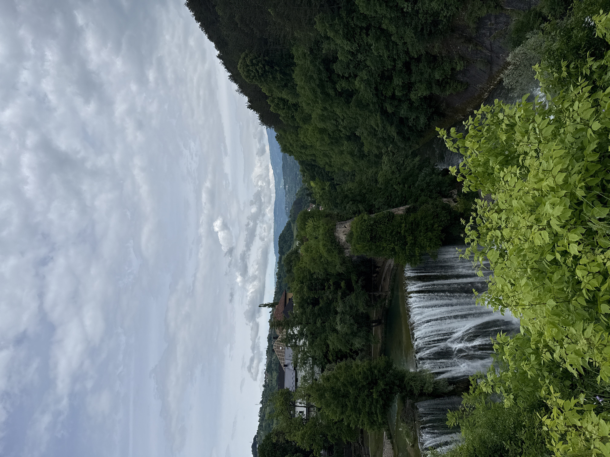 The image presents an elevated view of the Pliva Waterfall in Jajce, Bosnia and Herzegovina, under an overcast sky. In the foreground, vibrant green deciduous foliage fills the lower portion of the frame, partially obscuring the base of the waterfall and the river continuing into a deep gorge. The prominent feature is a wide, multi-stream waterfall cascading vertically from a placid body of water into the rocky, tree-lined canyon below. Above the waterfall, an expanse of the Pliva River is visible, bordered by dense clusters of diverse green trees and several residential or commercial buildings. These structures, situated on a raised landform, display varying architectural styles, including one with exposed roof trusses indicative of construction or renovation. Beyond these immediate elements, densely forested hills and mountains extend into the background, receding into a light blue atmospheric haze. The upper approximately two-fifths of the image are dominated by a heavily clouded sky, featuring numerous grey and white stratocumulus formations. No human figures or discernible activity are present.