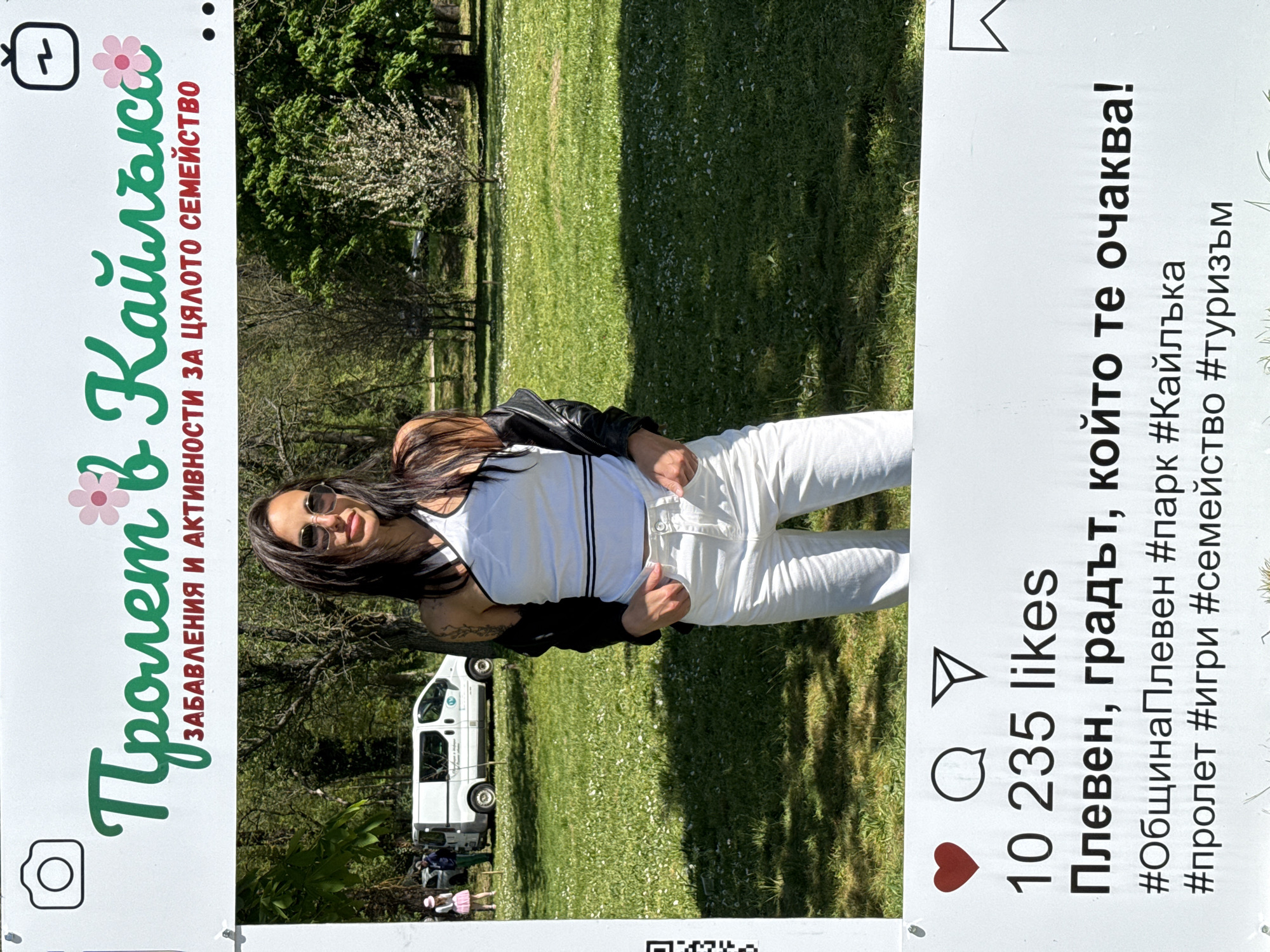 Here is a detailed description of the image:
**Scene:** The image captures a woman posing in front of an Instagram-style photo frame in what appears to be a public park. The frame is designed to look like a social media post and is located in the city of Pleven, Bulgaria. The park is likely the "Kaylaka" park, based on the text on the frame.
**People:**
* A woman is posing within the frame. She is wearing a white top with black stripes, white pants, and a black jacket. She has sunglasses on and is smiling.
* In the background, beyond the frame, there are other people visible, including some near a white van.
**Objects:**
* An Instagram-style photo frame with text in Bulgarian. The text includes phrases like "Spring in Kaylaka" (Пролет в Кайлъка), "Entertainment and activities for the whole family", "Pleven, the city that awaits you!", and hashtags such as #ОбщинаПлевен (Municipality of Pleven), #парк (park), #Кайлъка (Kaylaka), #пролет (spring), #игри (games), #семейство (family), #туризъм (tourism).
* A white van is parked in the background, partially visible behind the woman.
**Environment:**
* The setting is a grassy park area with trees.
* The image is taken during the daytime with natural light.
* The environment suggests a public park meant for recreation and family activities.
**Actions and Interactions:**
* The woman is posing for a picture, using the frame as a prop.
* The presence of other people in the background suggests that the park is open to the public and being used for leisure activities.
